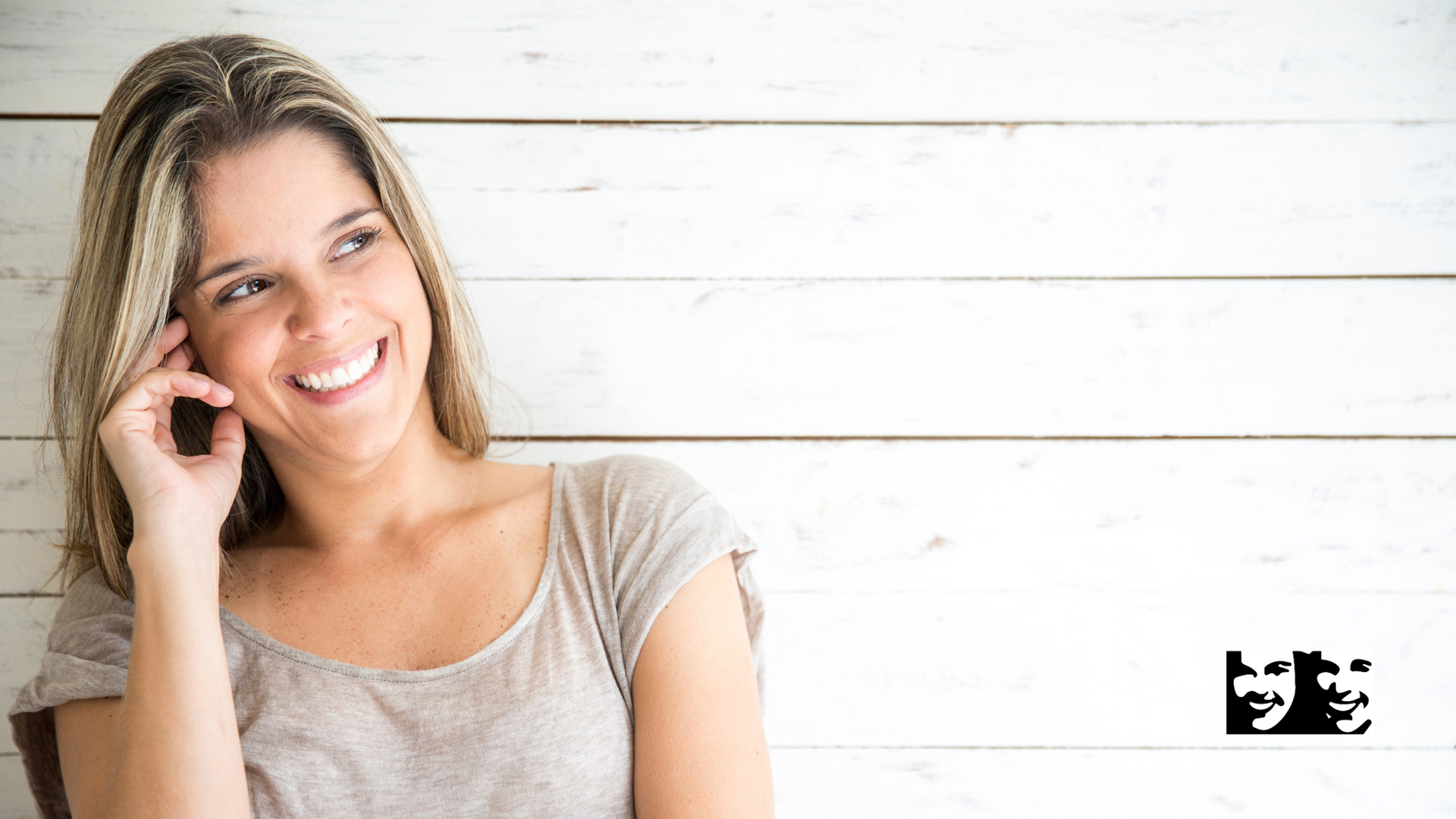 Woman with blonde hair smiling, leaning against a white wooden wall, hand near her face.