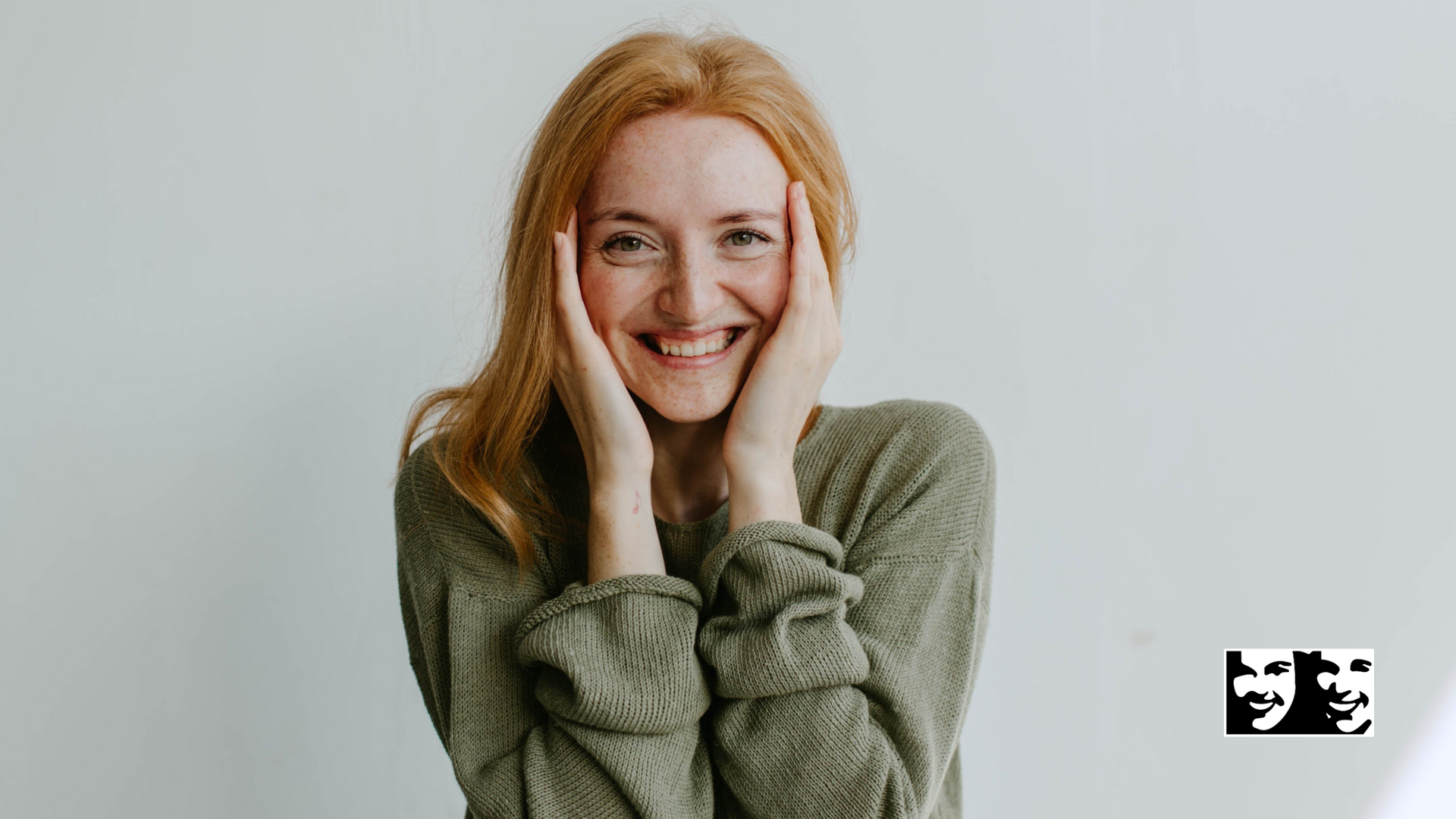 Woman with red hair, smiling, holding her cheeks, wearing a green sweater, against a white background.