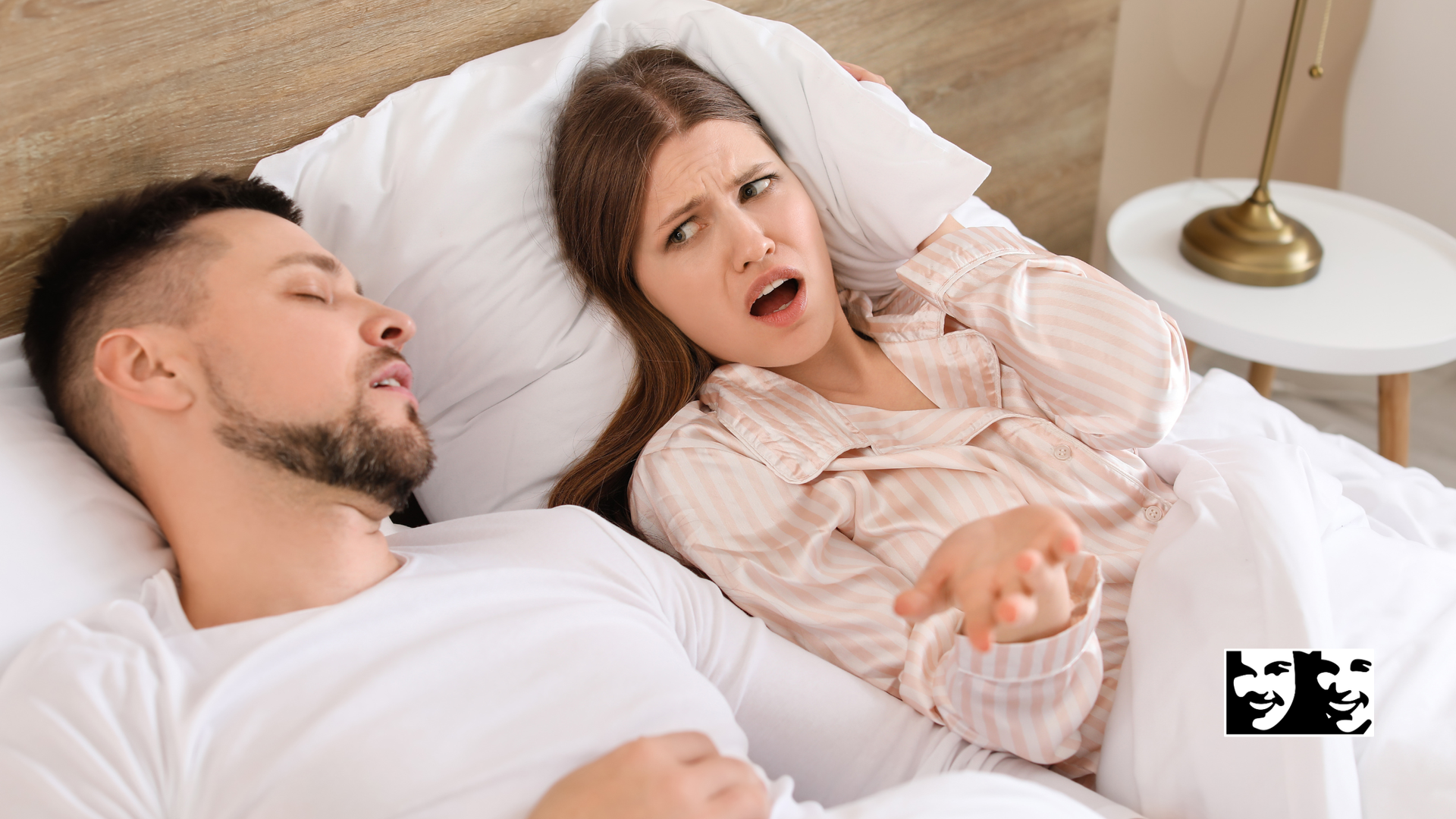 Couple lying in bed under white sheets, woman speaking while man sleeps beside her.