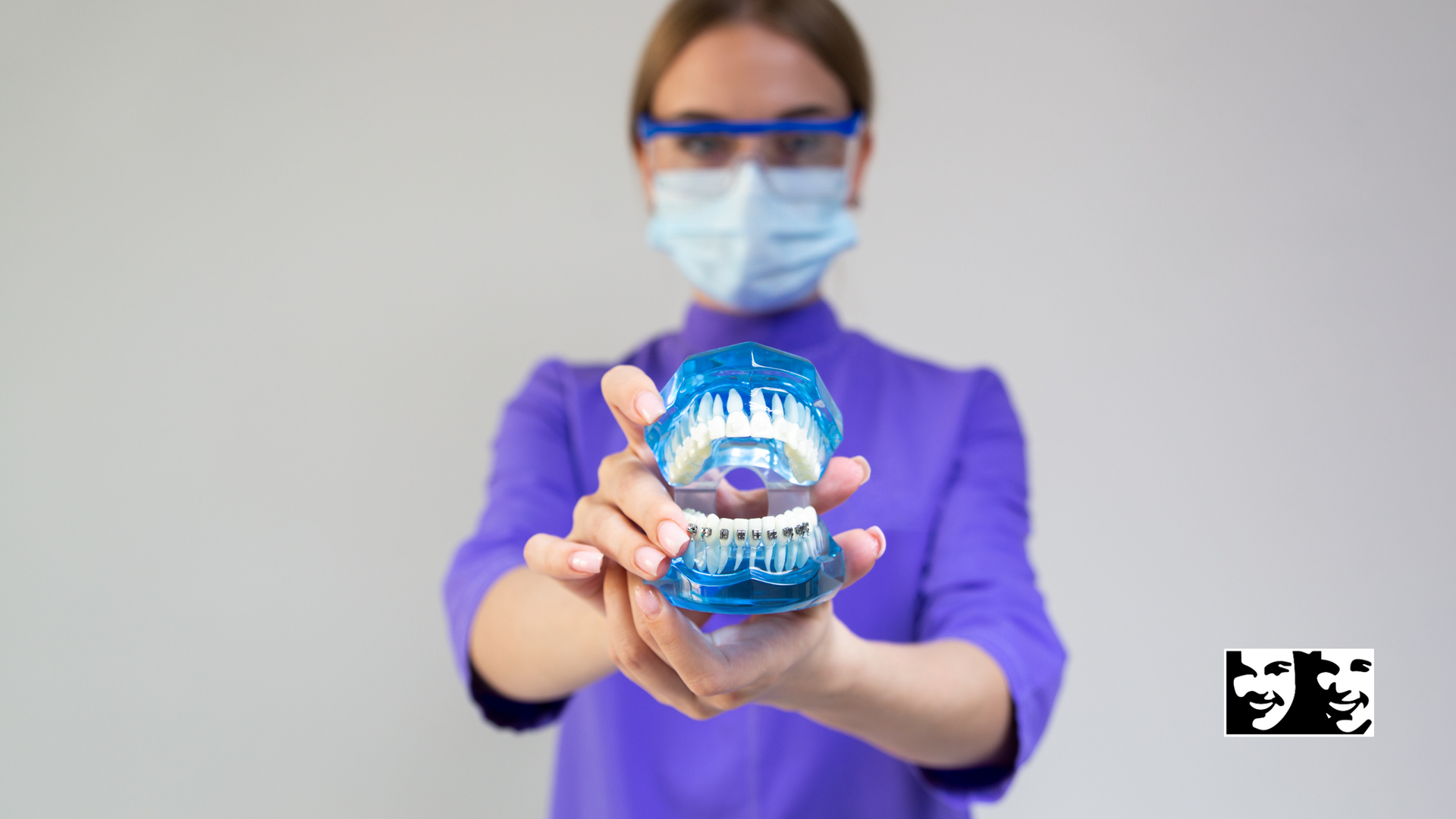 A dentist wearing a mask and glasses is holding a model of teeth.