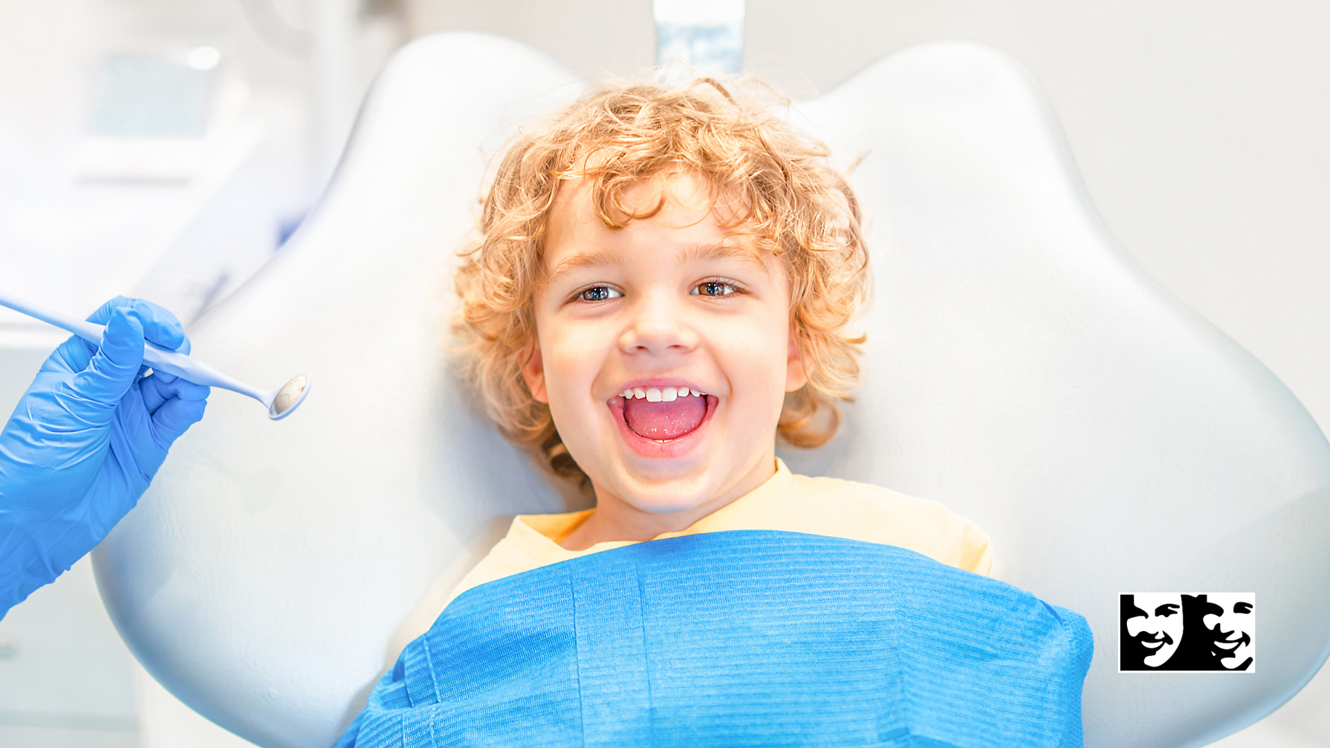 Boy with curly hair smiles wide in a dentist's chair as dentist holds tools.