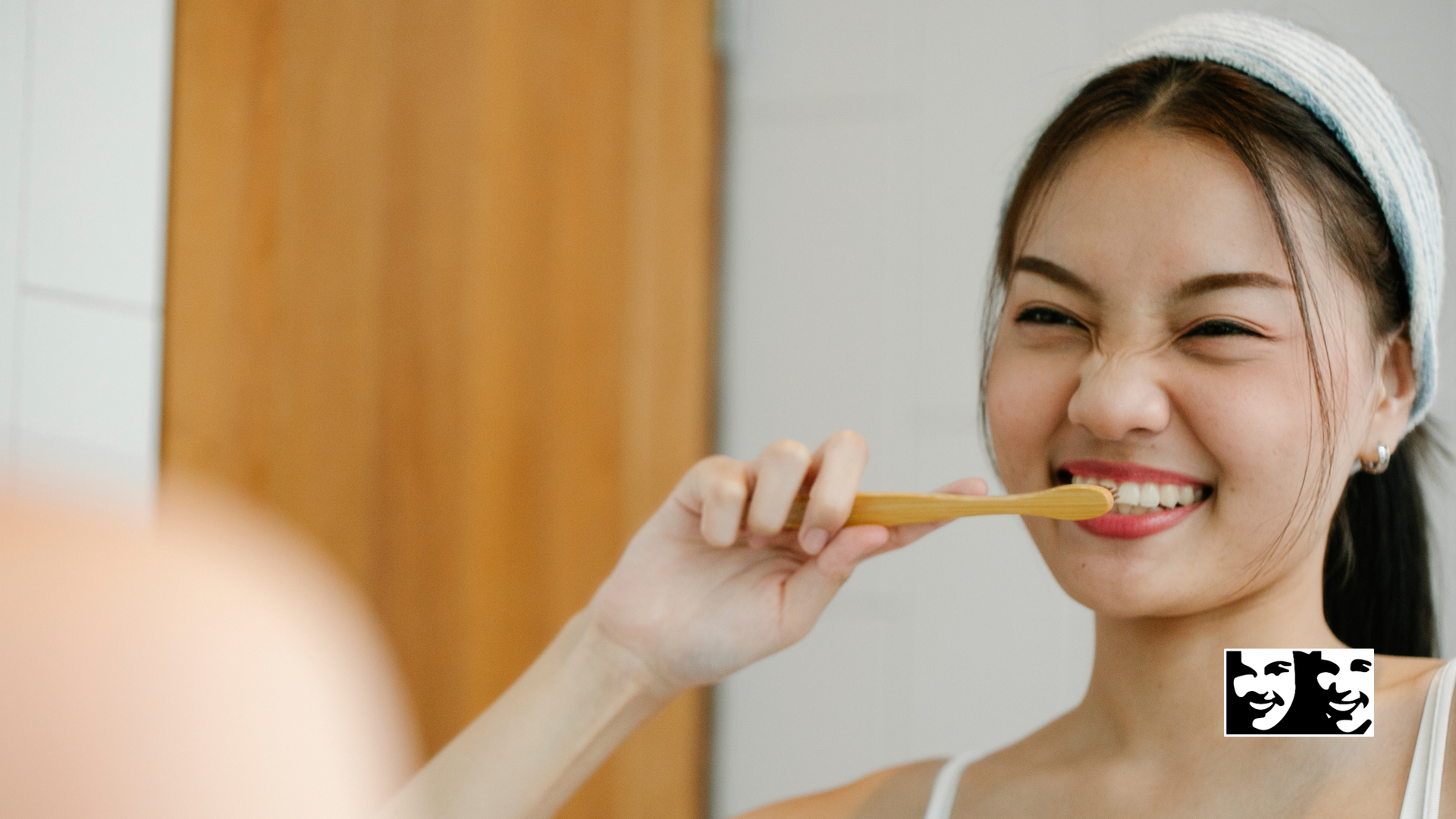 Woman brushing teeth in bathroom, smiling, wearing a headband, holding a wooden toothbrush.
