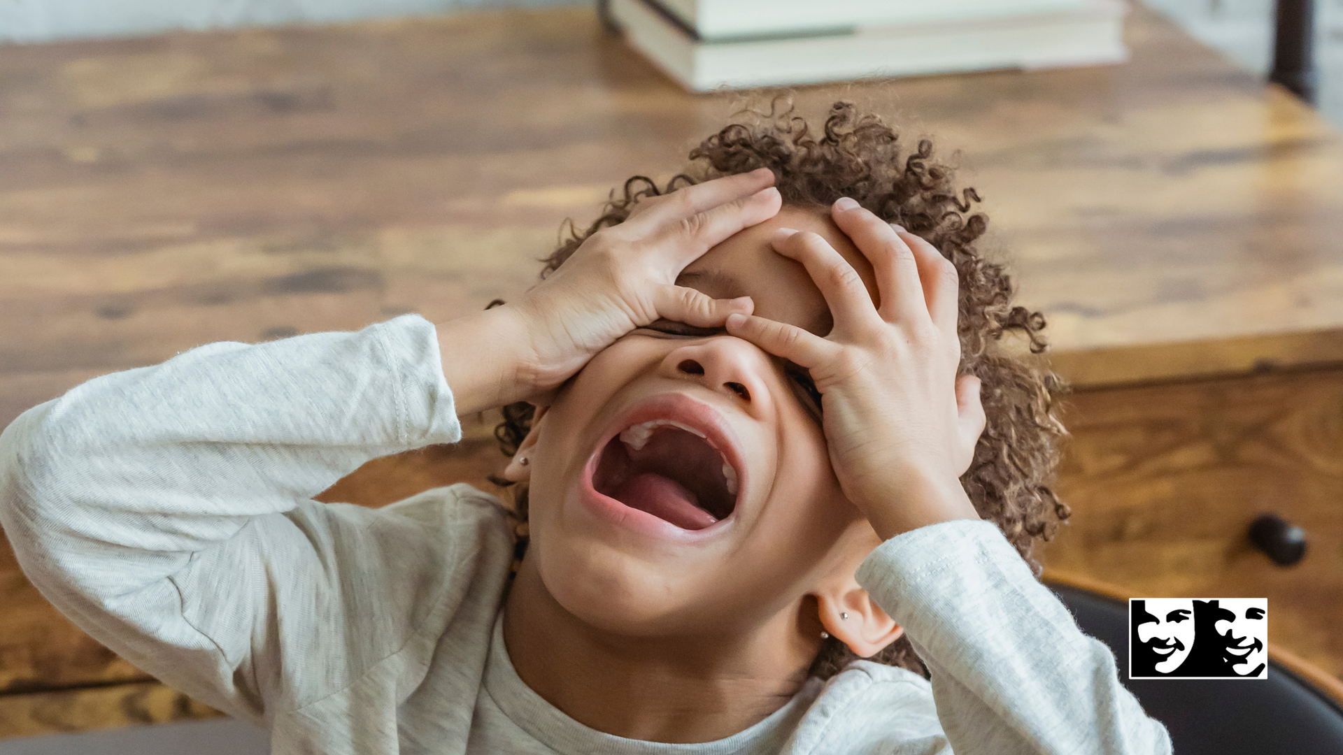 A young boy is crying with his mouth open and his hands on his head.