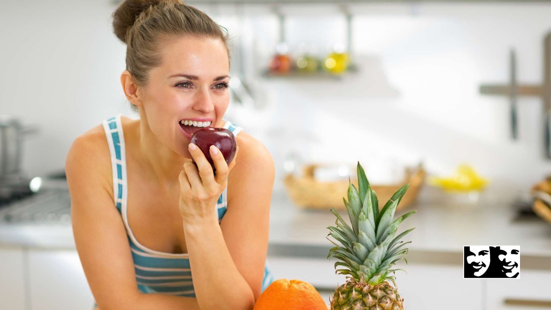 Woman eating an apple in a kitchen, with fruit on the counter, smiling.