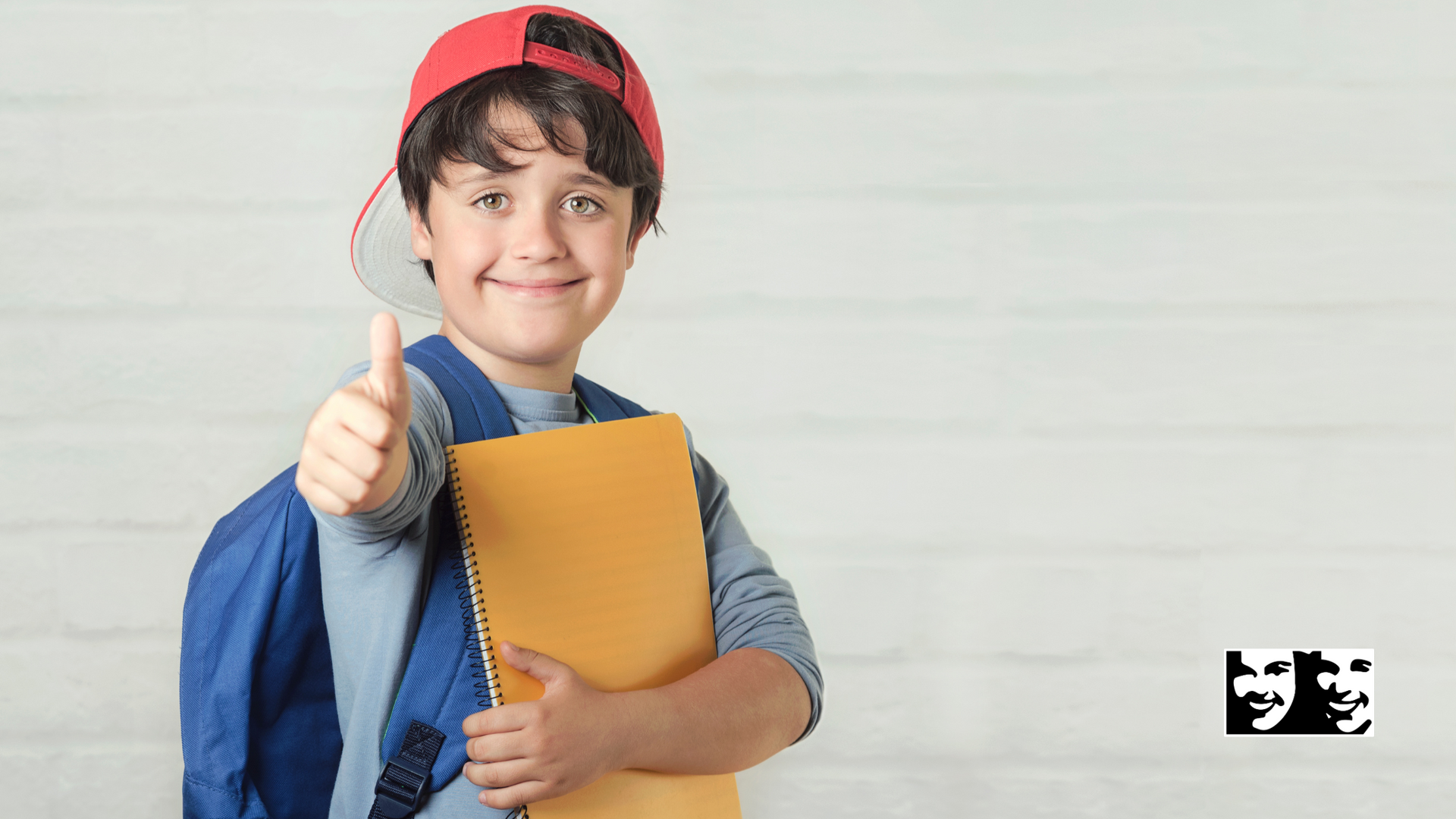 A young boy with a backpack is holding a notebook and giving a thumbs up.
