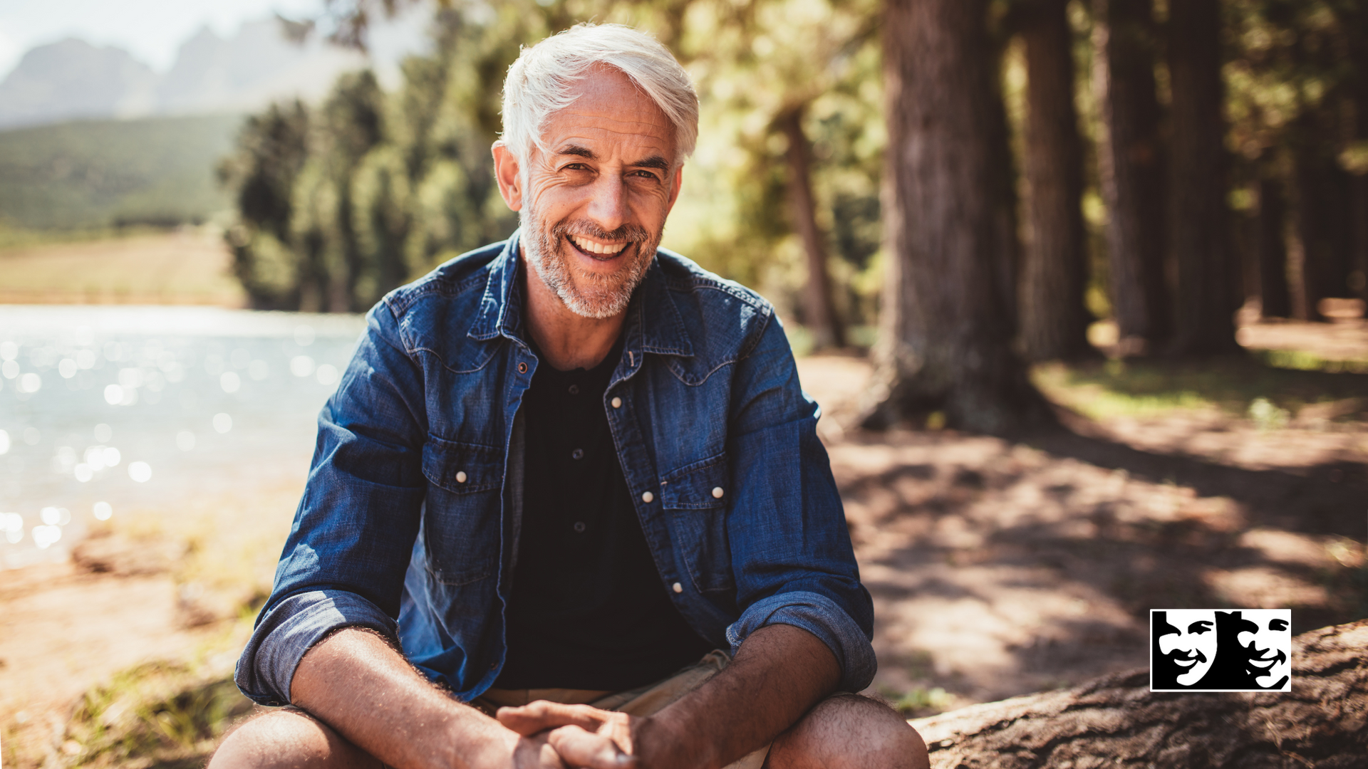 A man is sitting on a rock near a lake and smiling.