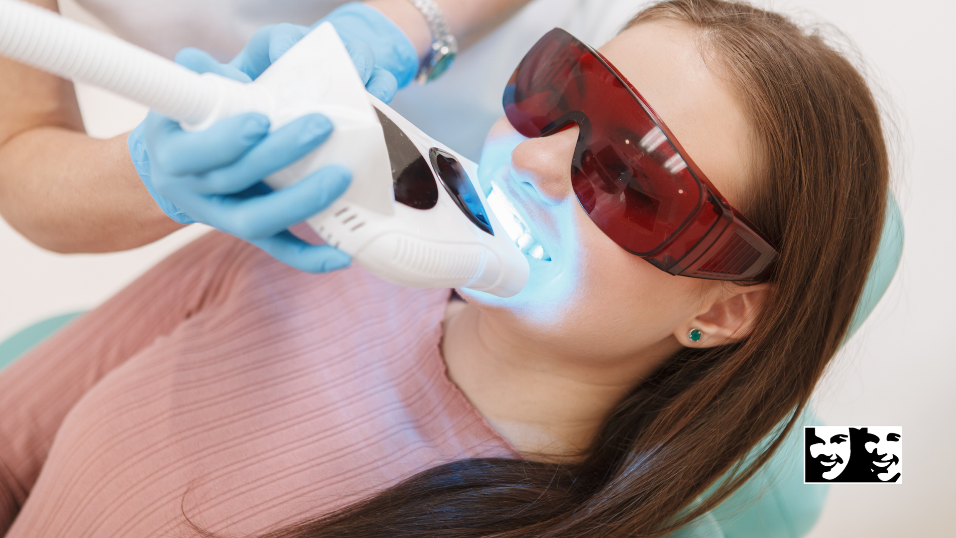 Woman receiving teeth whitening treatment; blue light directed at teeth; wearing protective glasses.