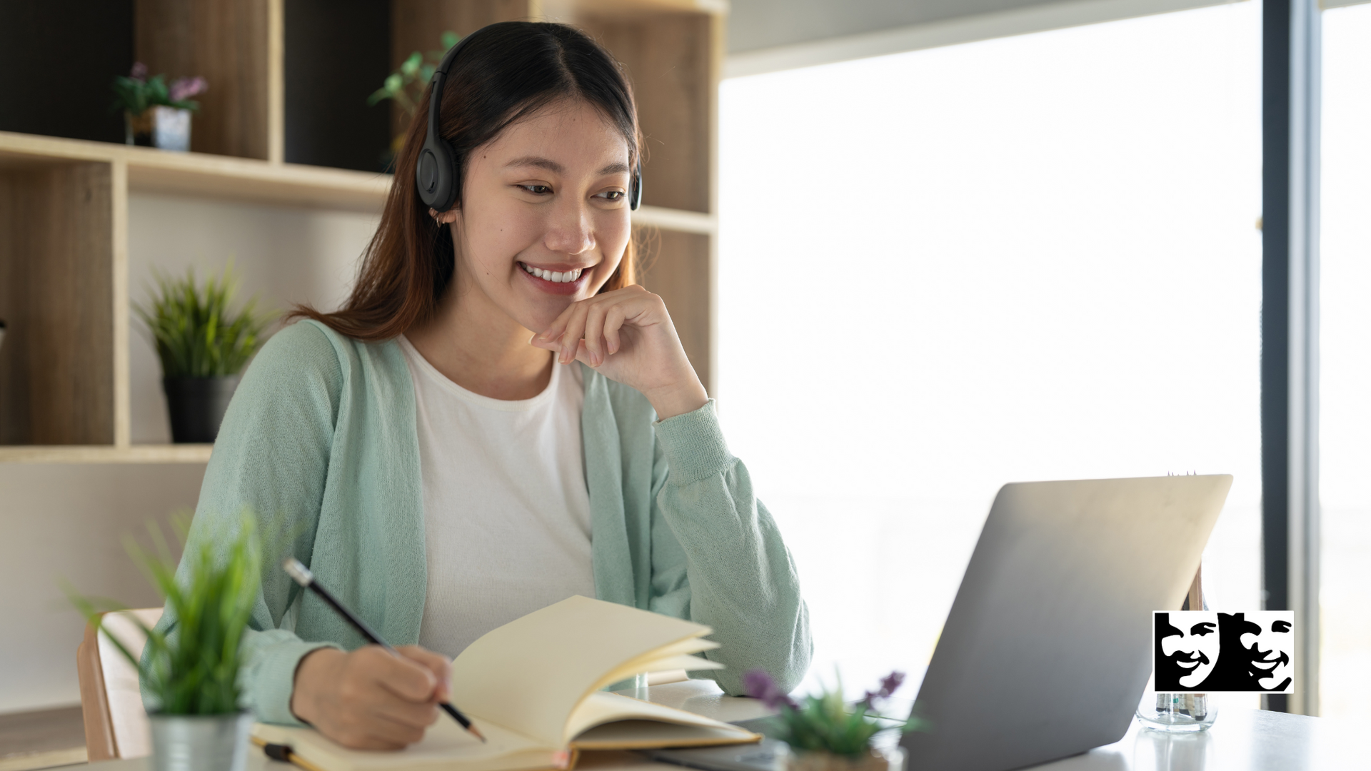 Woman with headphones takes notes while looking at a laptop, smiling, in a home office.