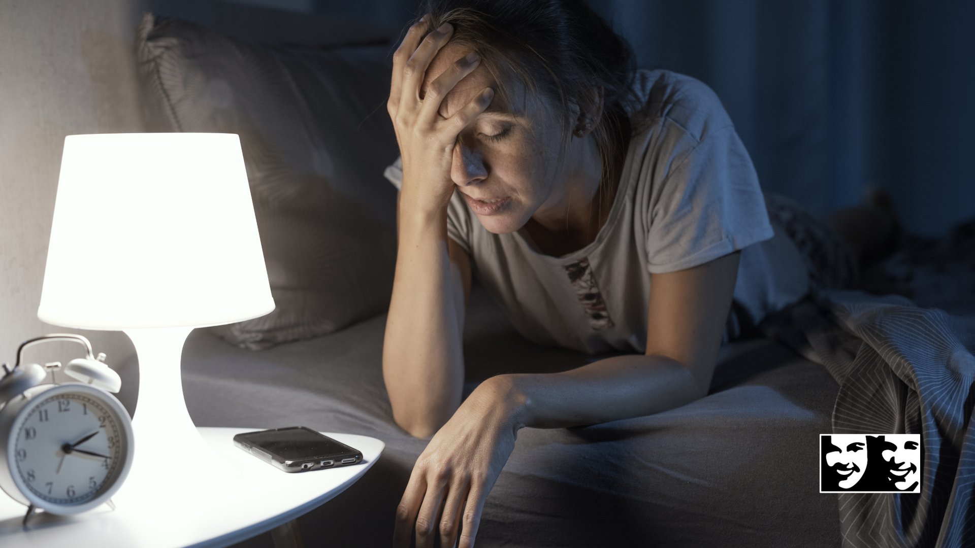 Tired person sitting by a lit bedside lamp, holding their head in a dark bedroom