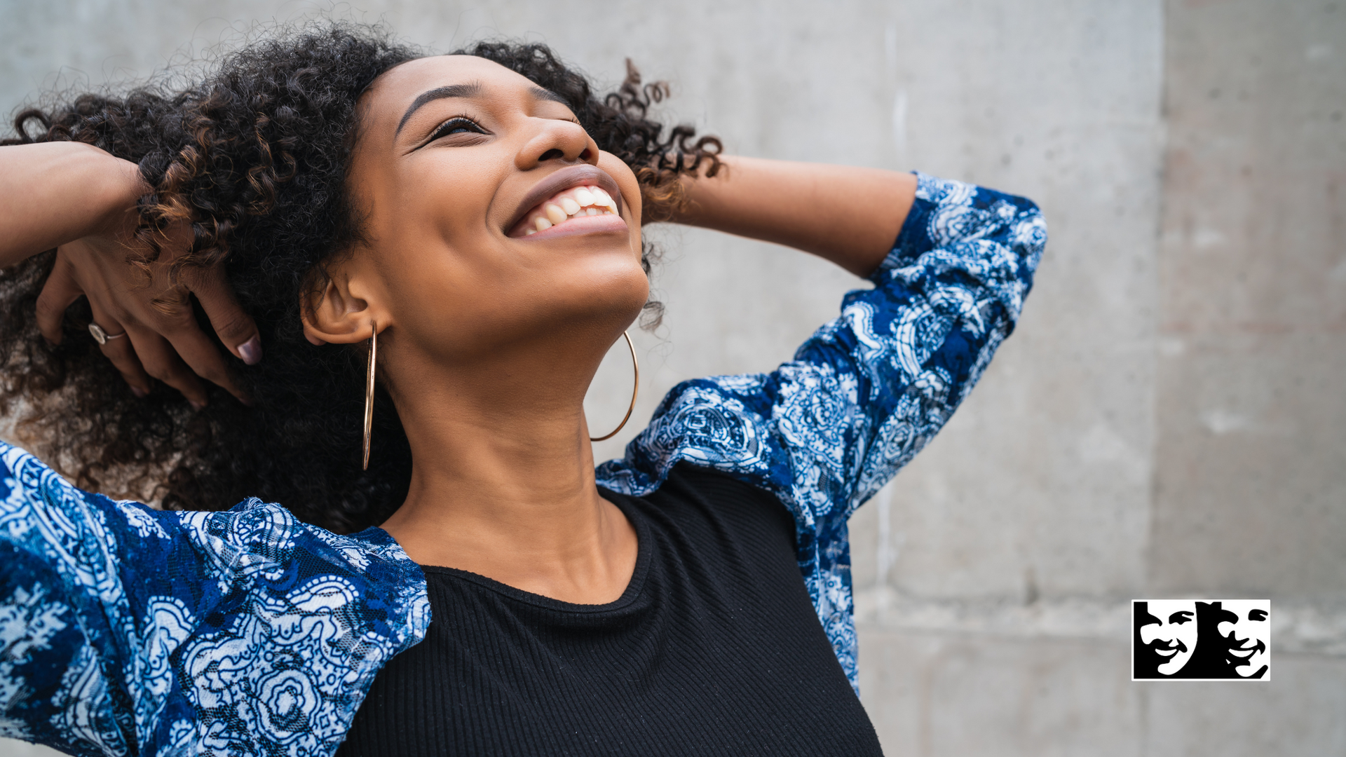 A woman with curly hair is smiling with her hands behind her head.