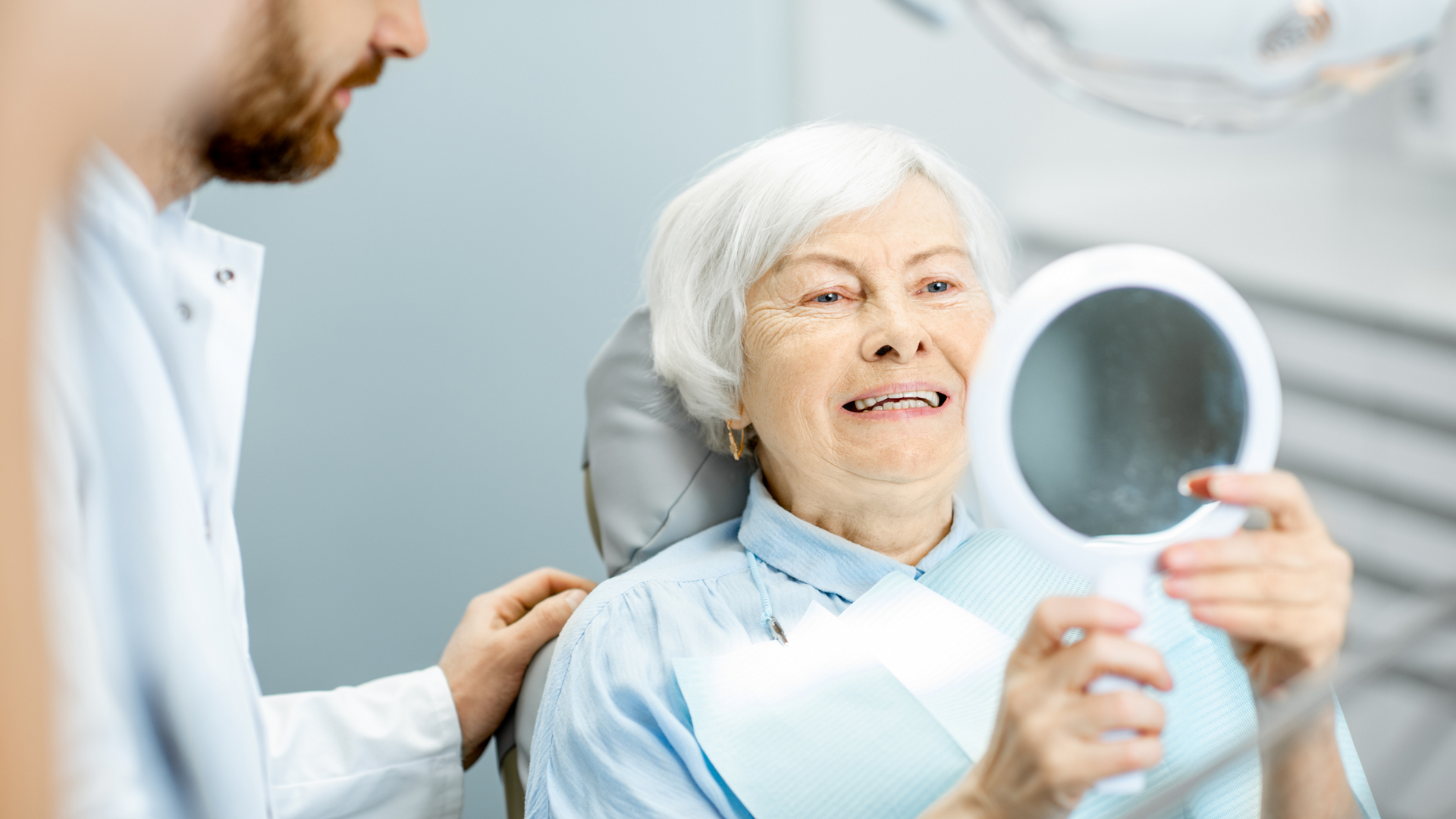 An elderly woman is looking at her teeth in a mirror.