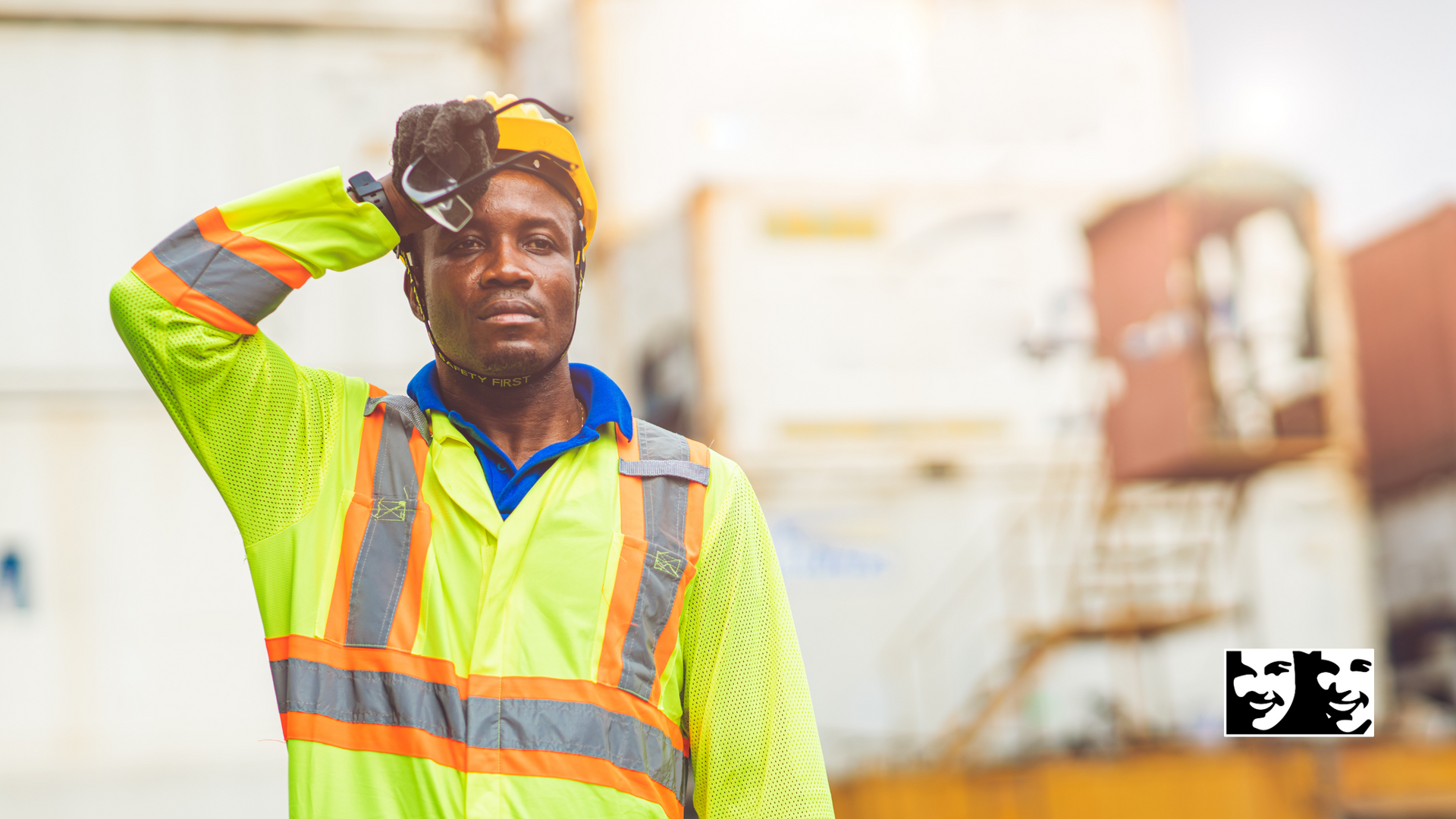 A construction worker is standing in front of a building holding his hand to his forehead.