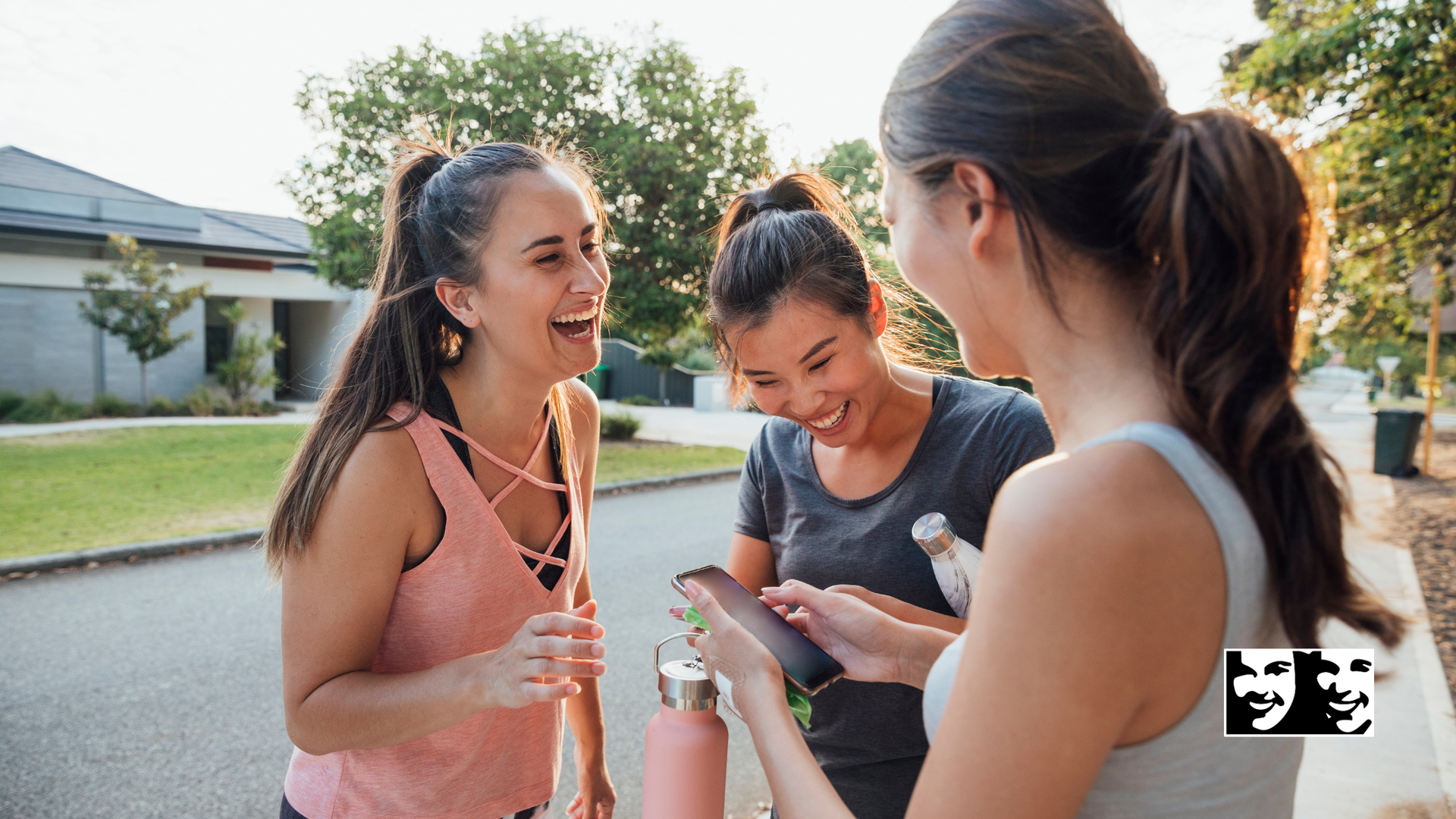 Three women laughing and looking at a phone on a sunny street.