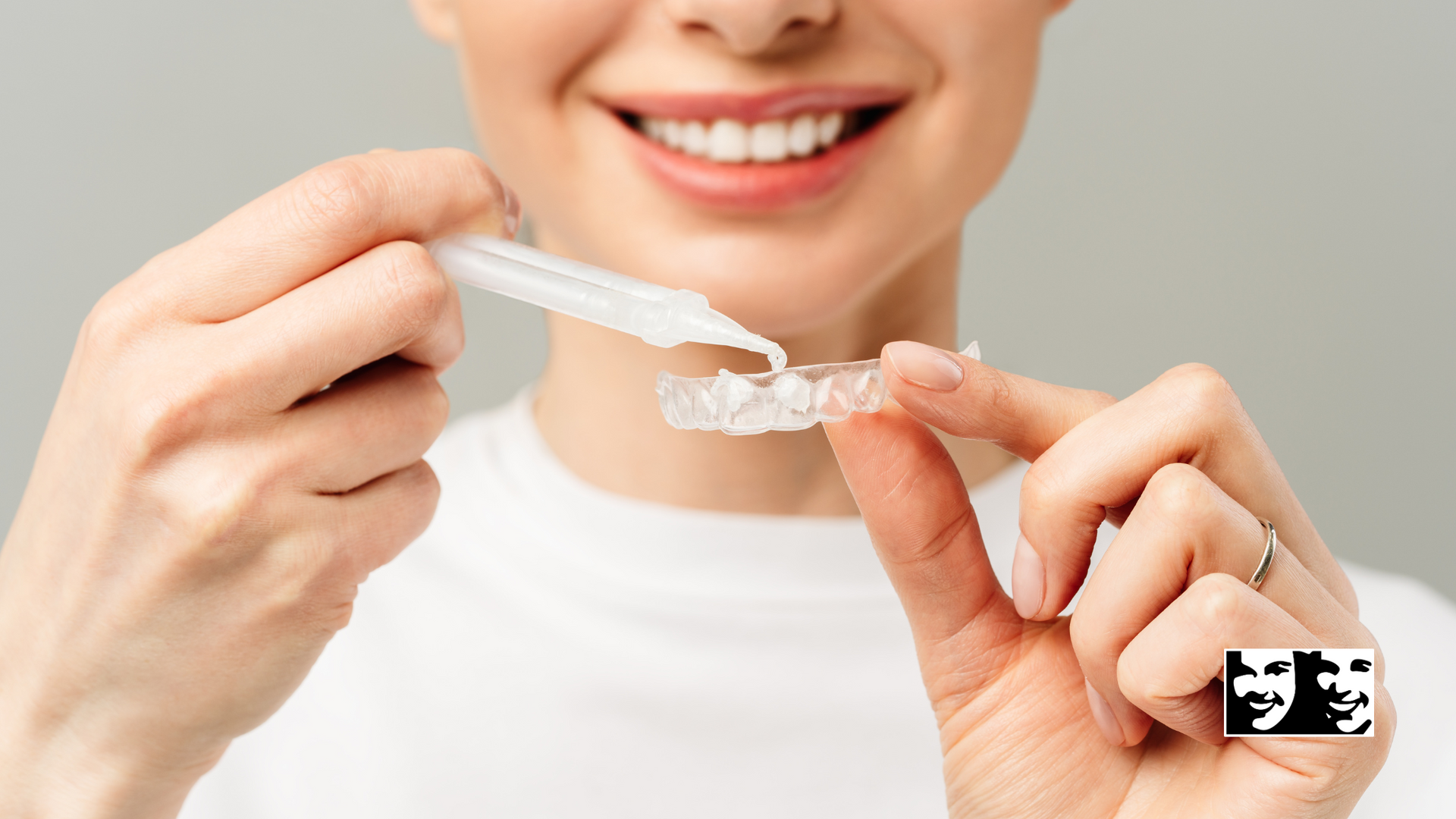 Woman applying whitening gel to a teeth whitening tray, smiling against a gray background.
