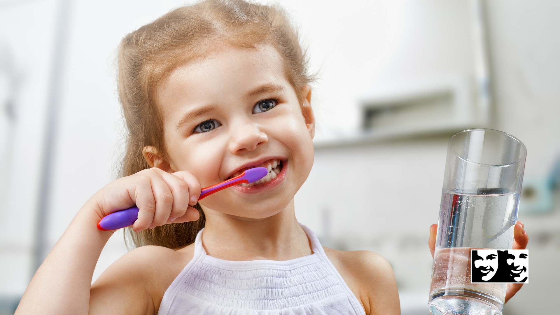 A little girl is brushing her teeth while holding a glass of water.