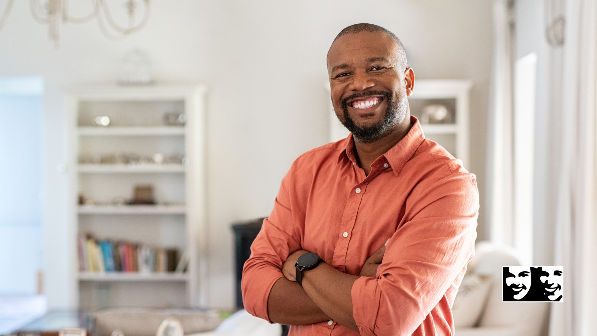Smiling Black man with arms crossed, wearing orange shirt. Standing indoors near windows and furniture.