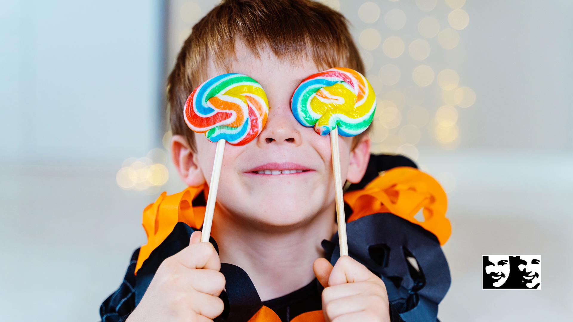 Boy holding two colorful lollipops up to his eyes, with a Halloween decoration around his neck.