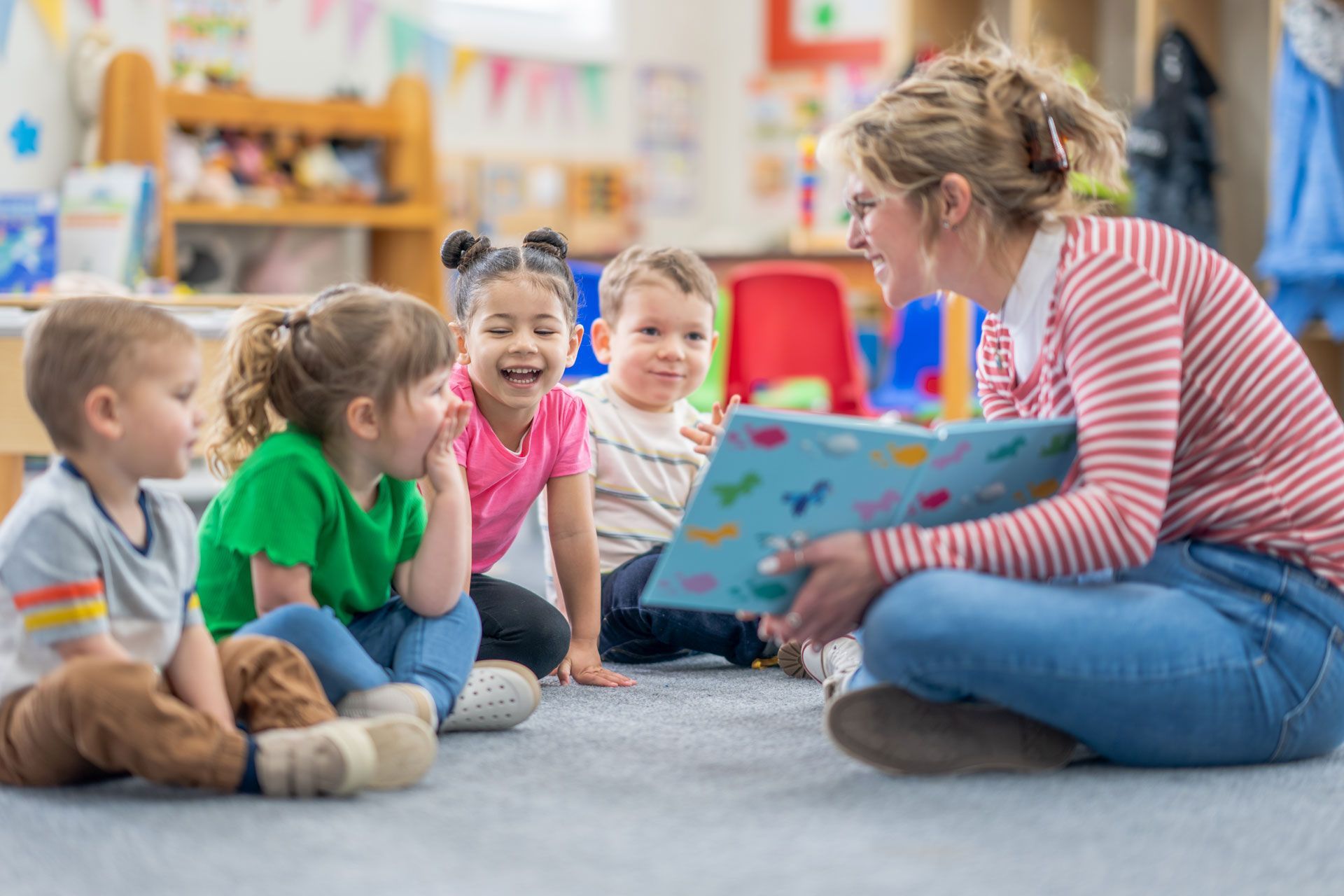 A preschool teacher sits on the floor with a small group of students as she reads them a book. A preschool teacher sits on the floor with a small group of students as she reads them a book.