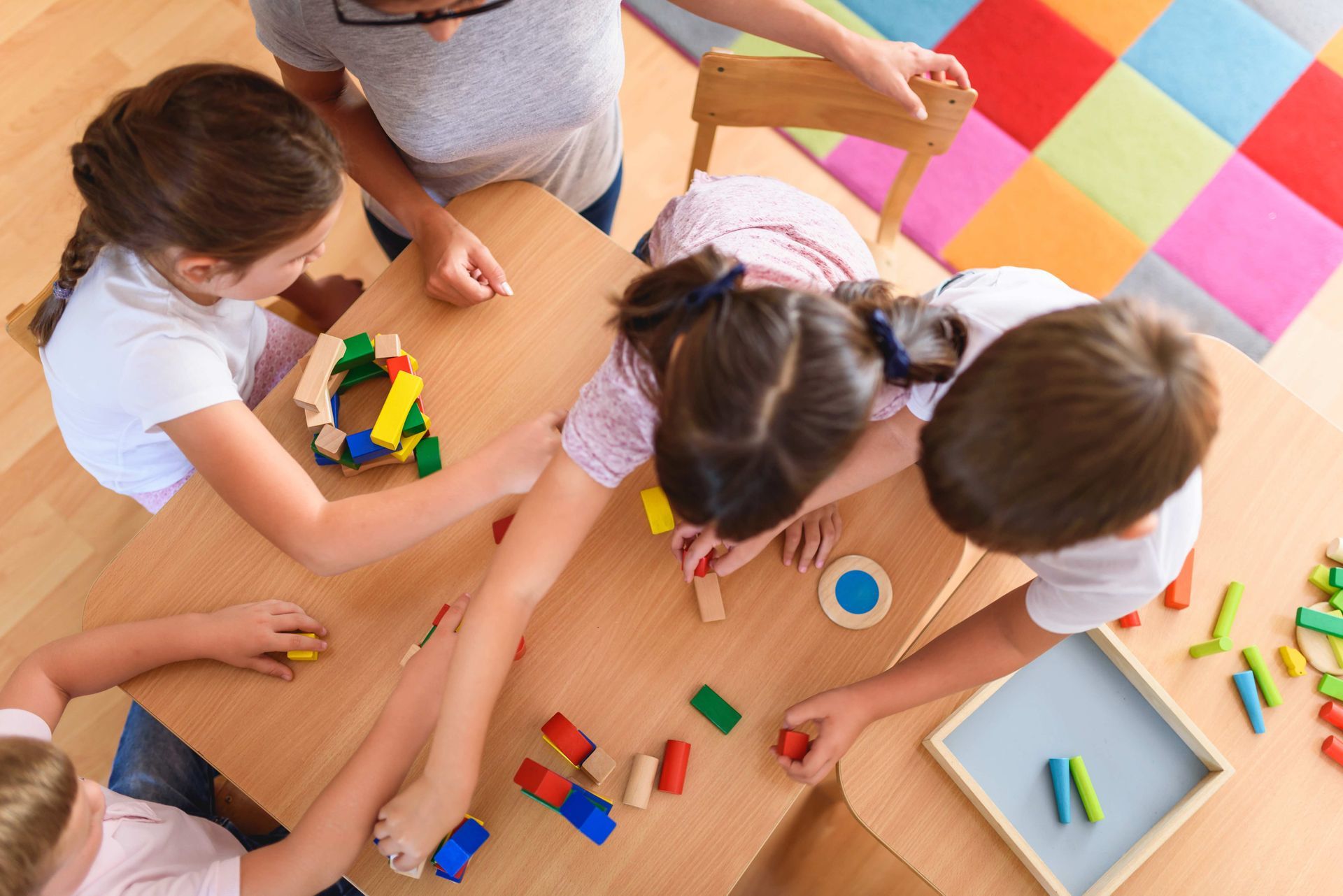 Children playing with colorful wooden blocks at a table while a teacher supervises Children playing with colorful wooden blocks at a table while a teacher supervises