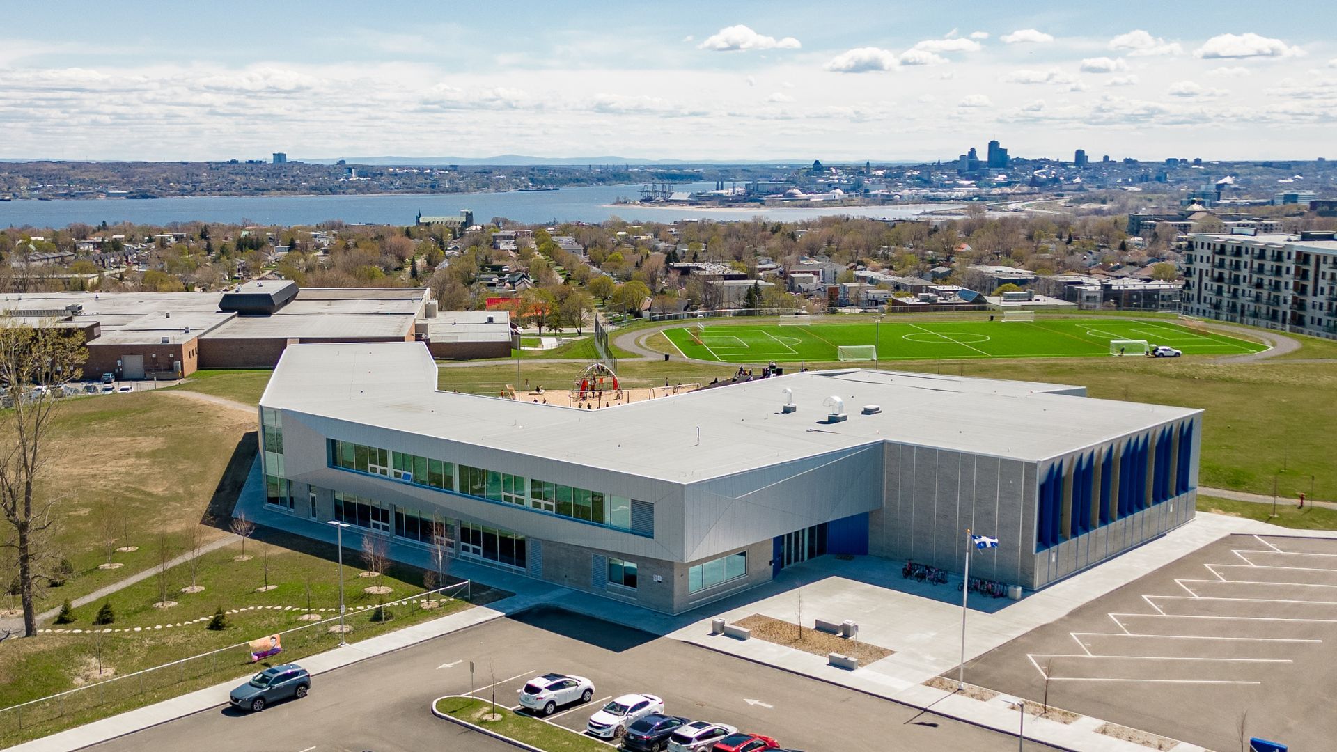 Vue aérienne d'un bâtiment scolaire moderne près d'un champ et de l'eau, avec des accents bleus et des voitures dans un parking.