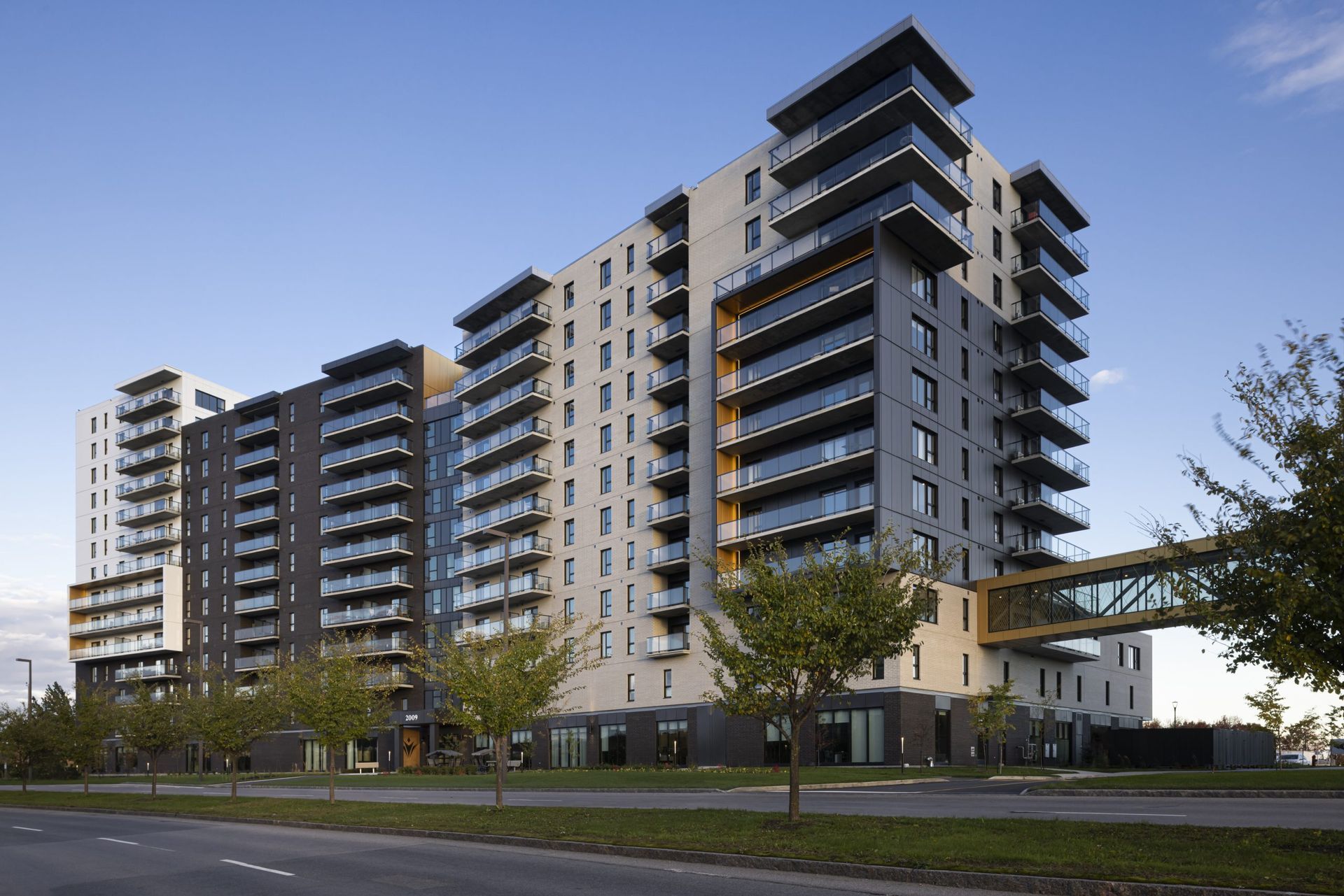Immeuble d'appartements moderne à plusieurs étages avec balcons en verre, pont de liaison et ciel bleu.