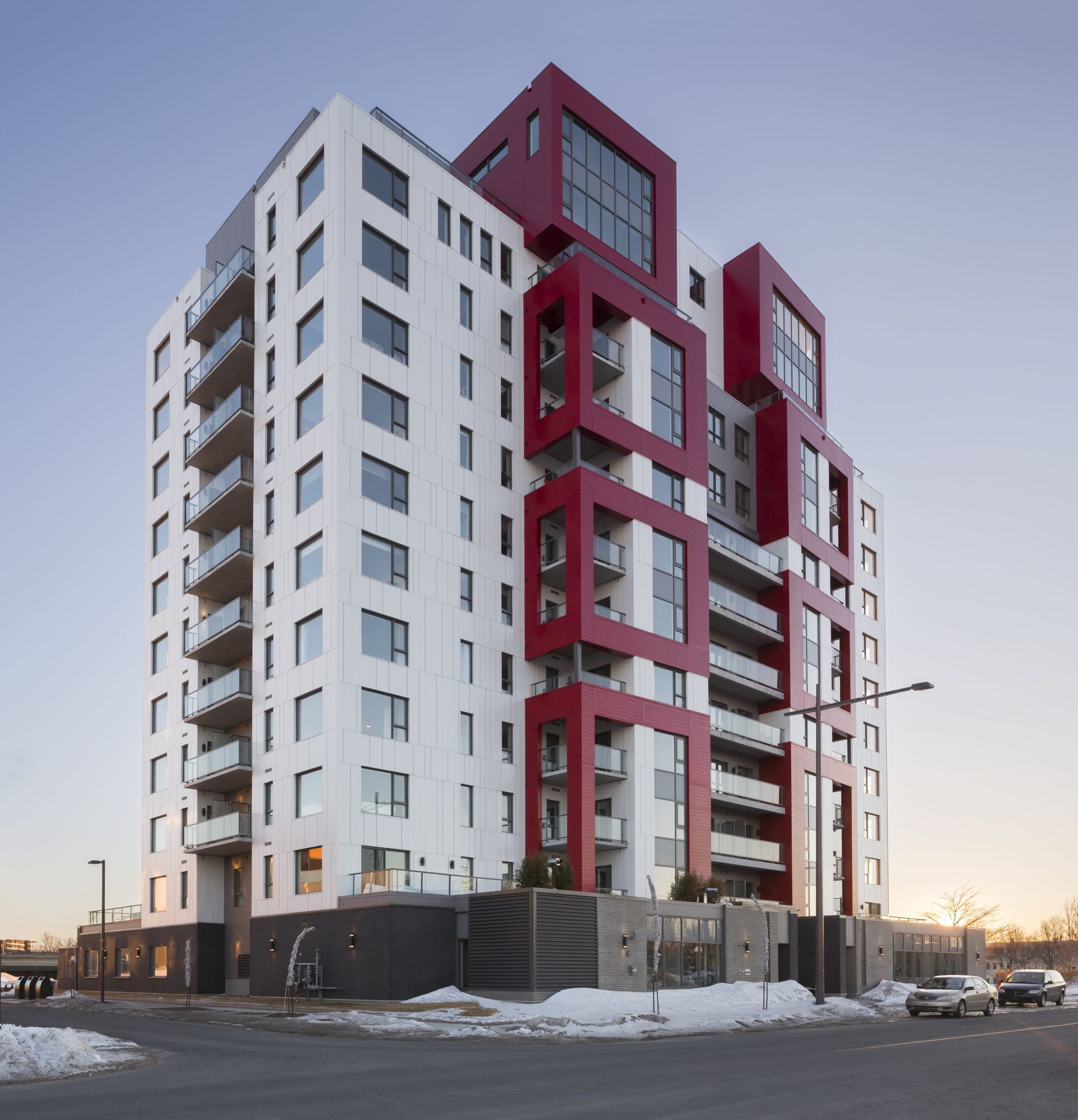 Immeuble d'appartements moderne de grande hauteur avec façade blanche, touches de rouge et balcons vitrés. Situé dans une rue enneigée.