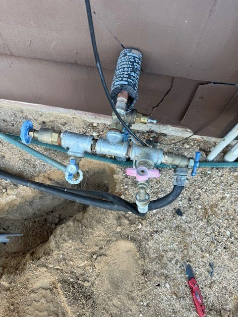 Outdoor plumbing manifold with blue and pink shutoff valves and black hoses beside a wall and dirt ground