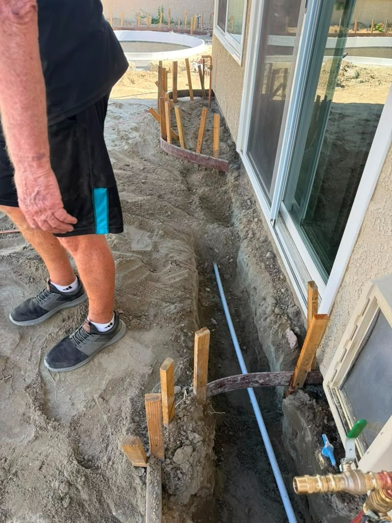 Worker beside house foundation trench with exposed pipes and stakes in sandy ground