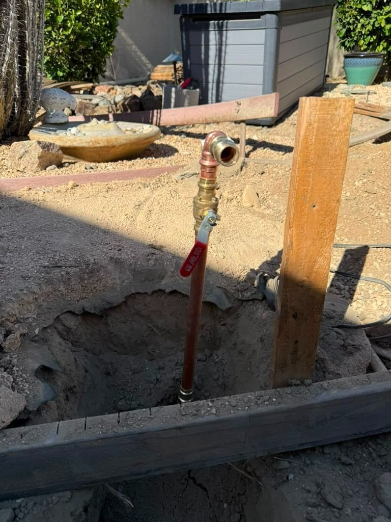 Outdoor construction scene with a red water pipe and brass valve beside a wooden post and gravel ground