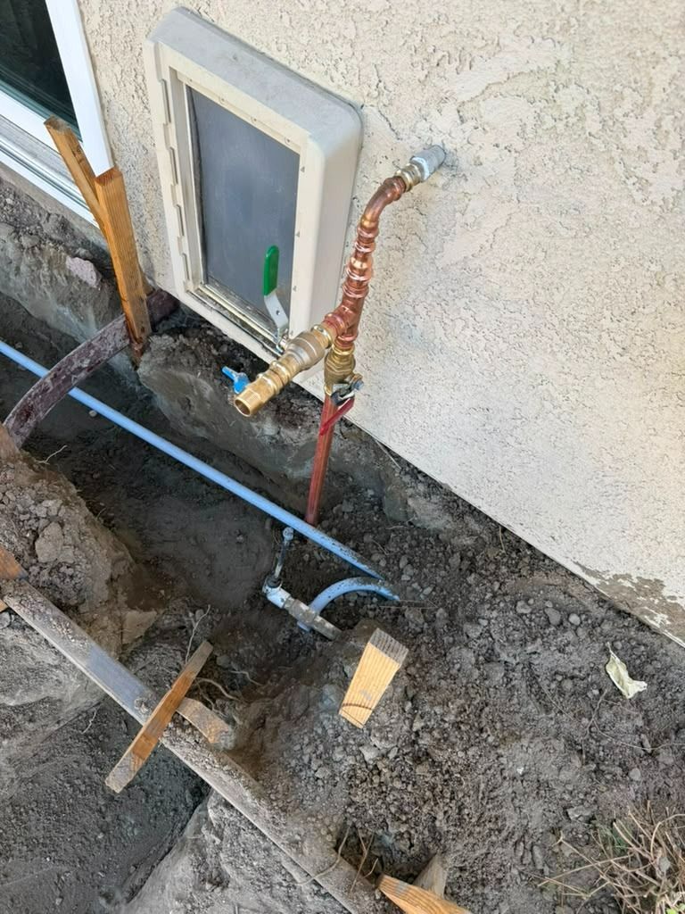 Outdoor utility trench beside a house wall, with exposed pipes, cables, and a small access panel