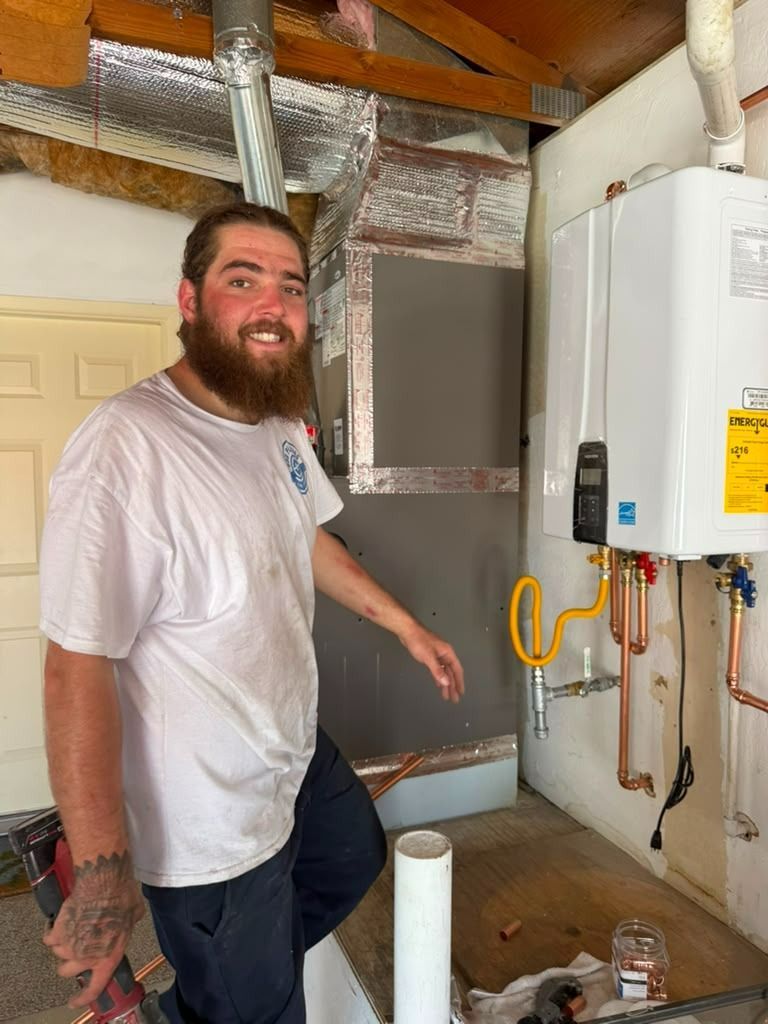 Man standing in a basement beside a wall-mounted water heater and exposed ductwork, smiling and gesturing.