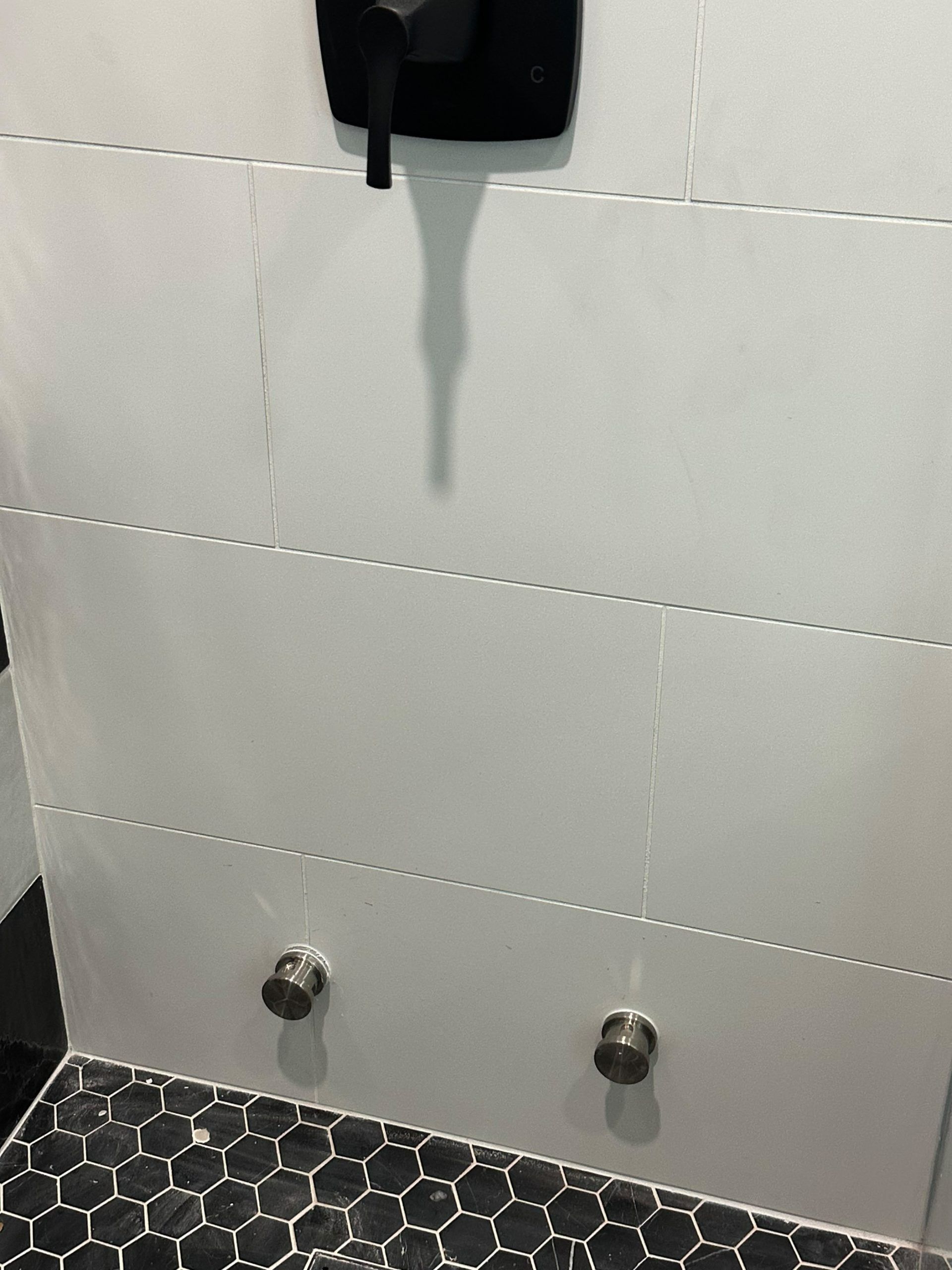 Tiled shower wall with a black shower fixture and two metal knobs above a hexagon-tile floor.