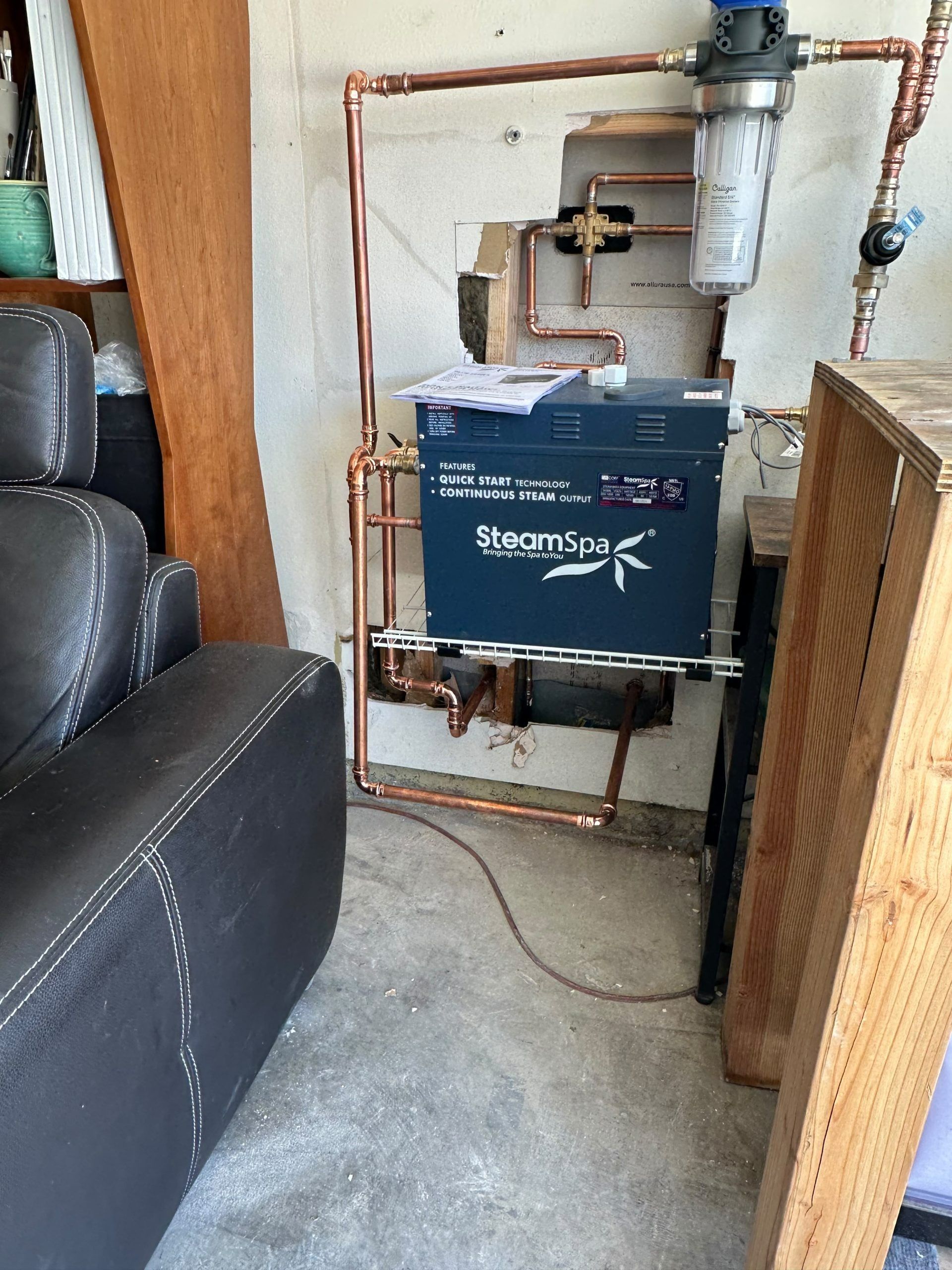 Boiler and exposed copper pipes in a utility closet beside a couch and wood railing