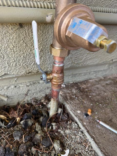 Close-up of a brass gas regulator and copper pipe beside a stucco wall, with an exposed line in dirt and debris.