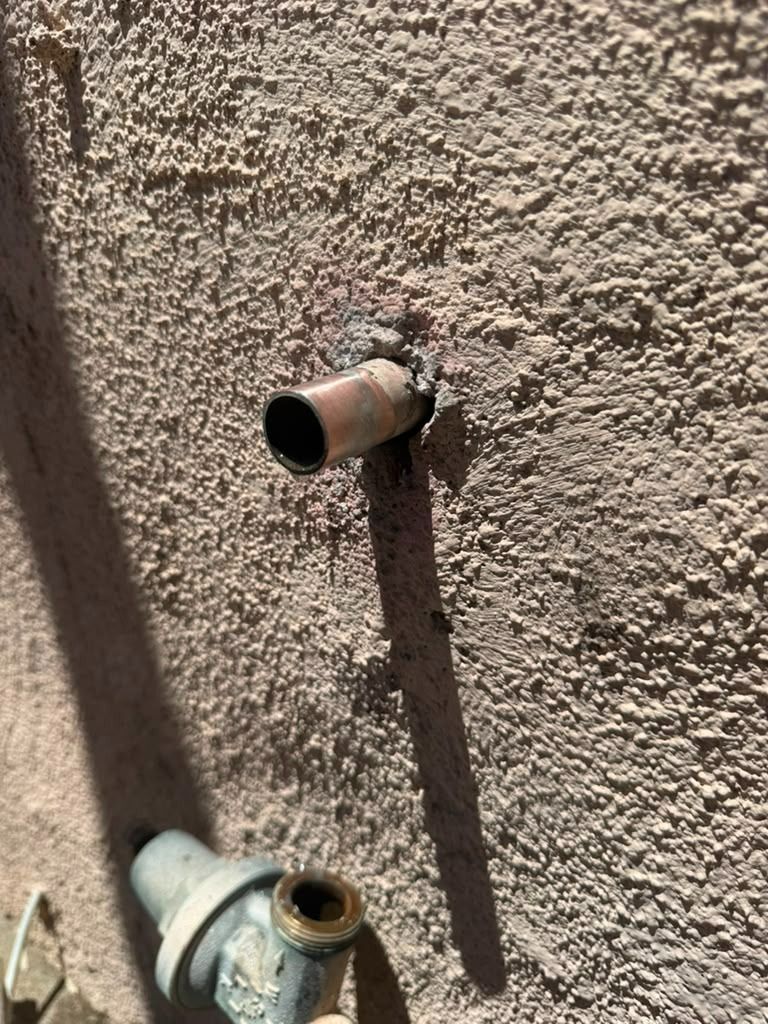 Two wall-mounted metal pipes protruding from a textured stucco wall, with a longer shadow cast below.