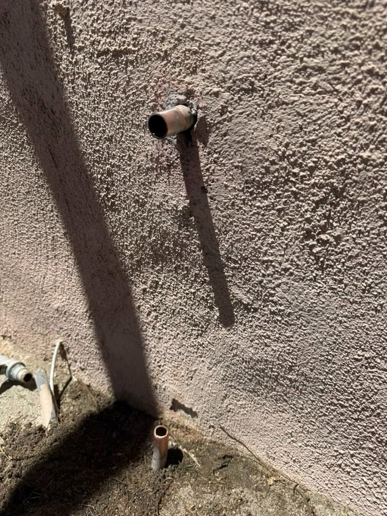 Outdoor stucco wall with two exposed pipes casting long shadows near the ground