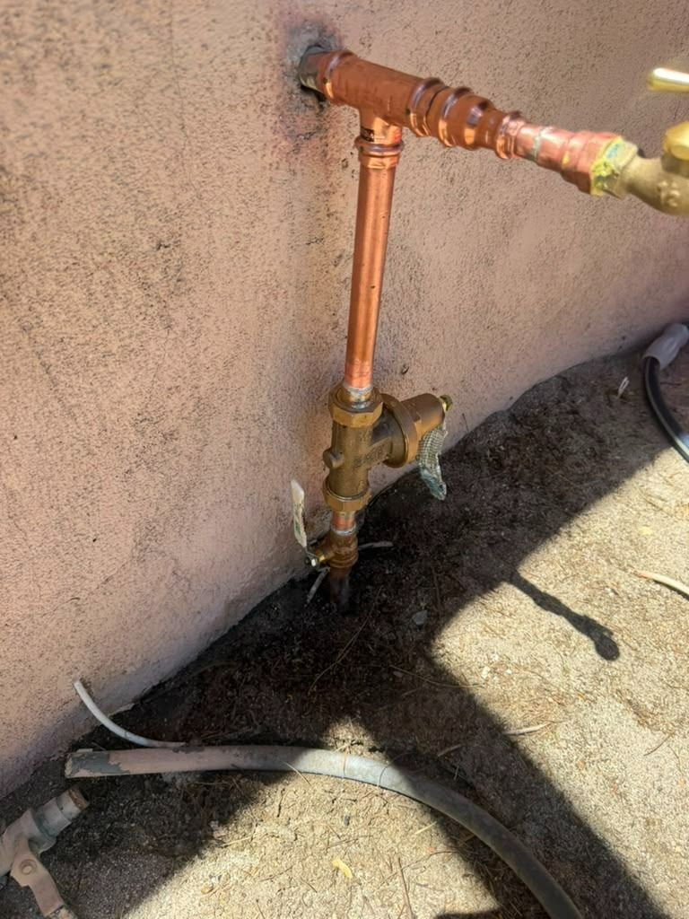 Copper pipe and brass valve mounted on an outdoor stucco wall beside a hose on the ground