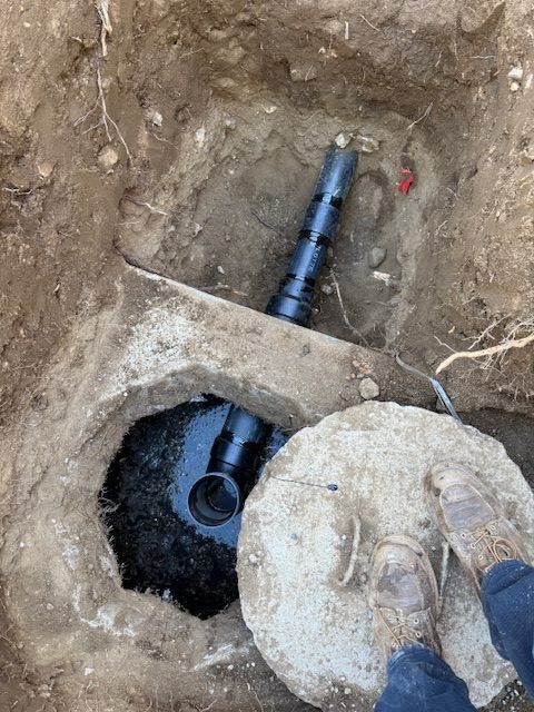Black pipe and concrete utility access in a dirt hole, with a round lid on the right.