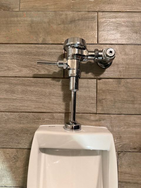 Chrome flush valve above white urinal against wood-look tiled wall