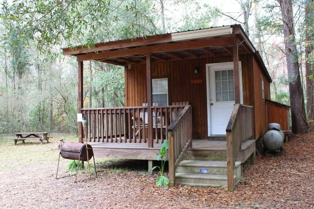 Small rustic wooden cabin with a porch in a wooded clearing, with a grill in front.