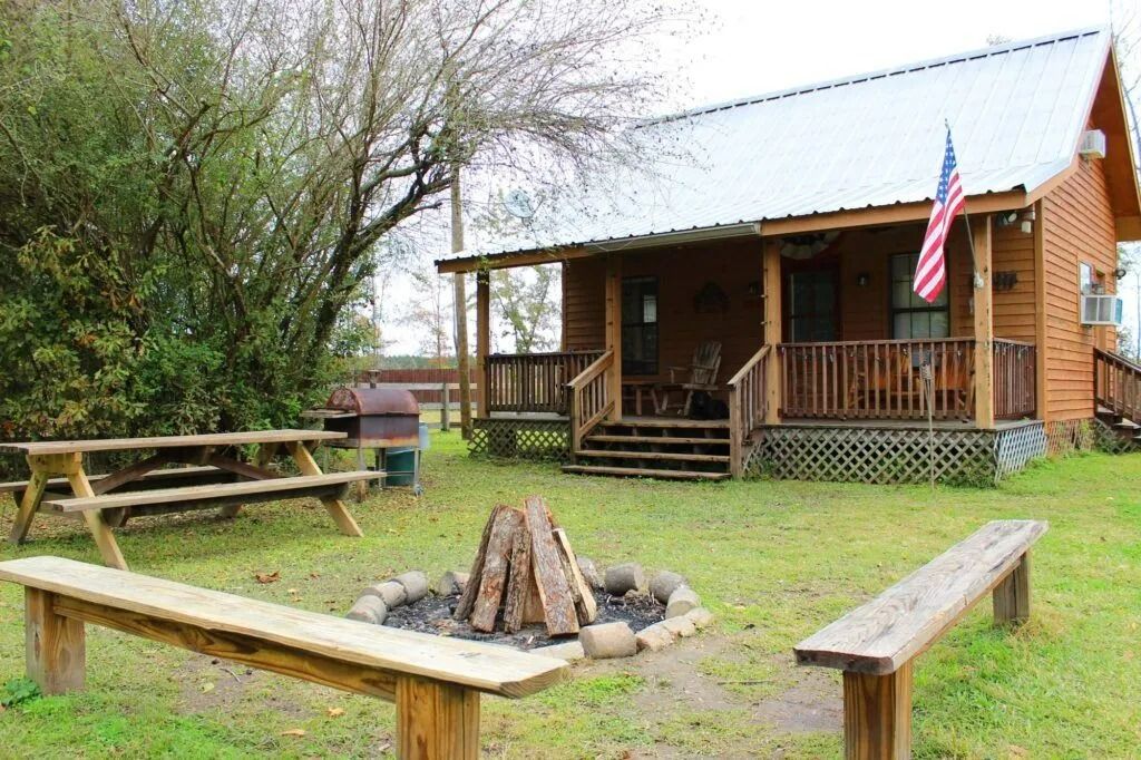 Rustic cabin with porch and American flag, picnic tables, and a fire pit in a grassy yard