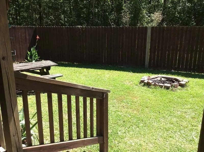 Sunny backyard with green lawn, wooden deck railing, fence, and a fire pit in the center.