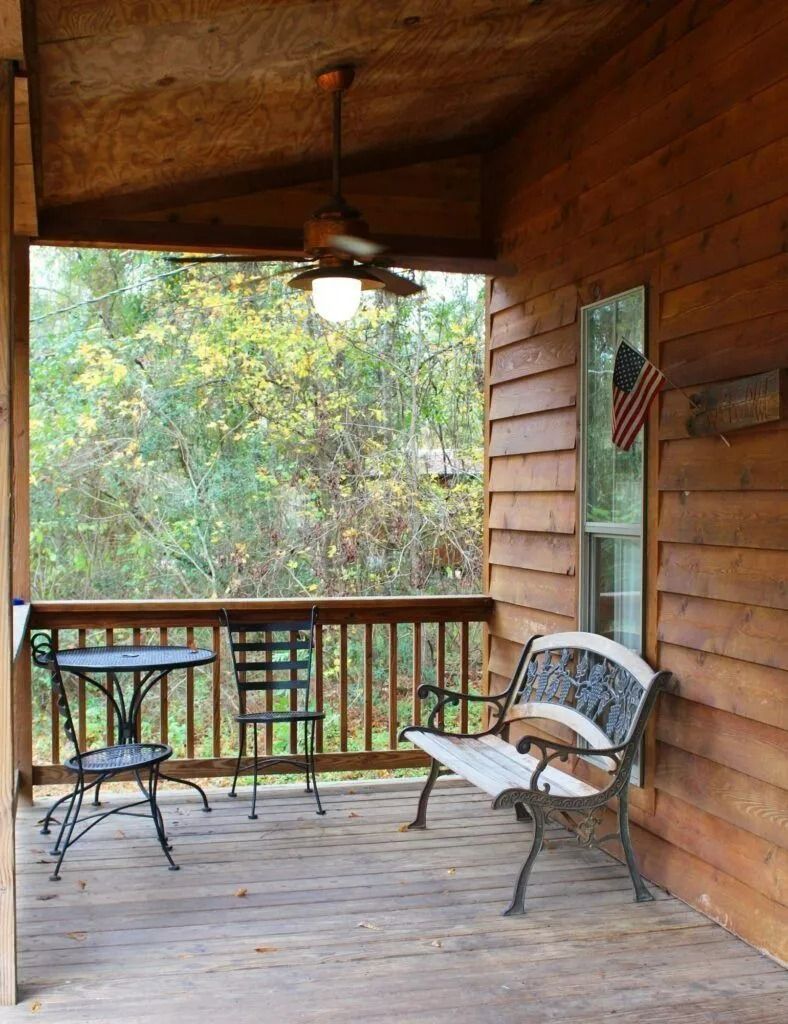 Wooden porch with bench, table, ceiling fan, and view of trees beyond the railing.
