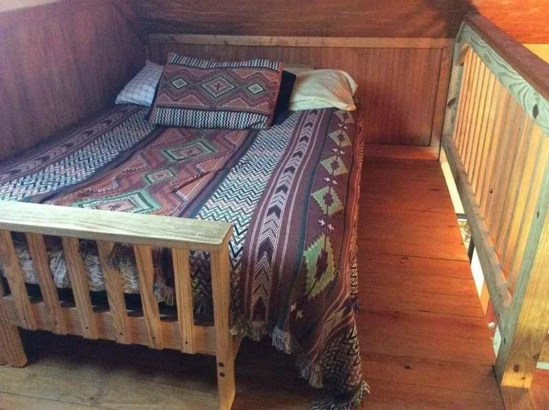 Small wooden bed in a cozy room, made with a patterned blanket and pillows near a sunlit window.