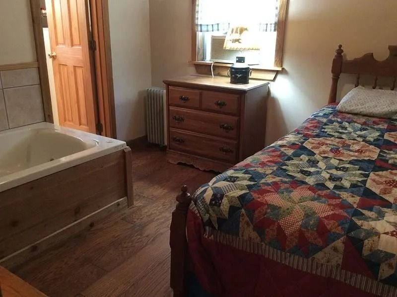Bedroom with wooden bed, quilt, dresser by window, and bathtub area visible to the left.