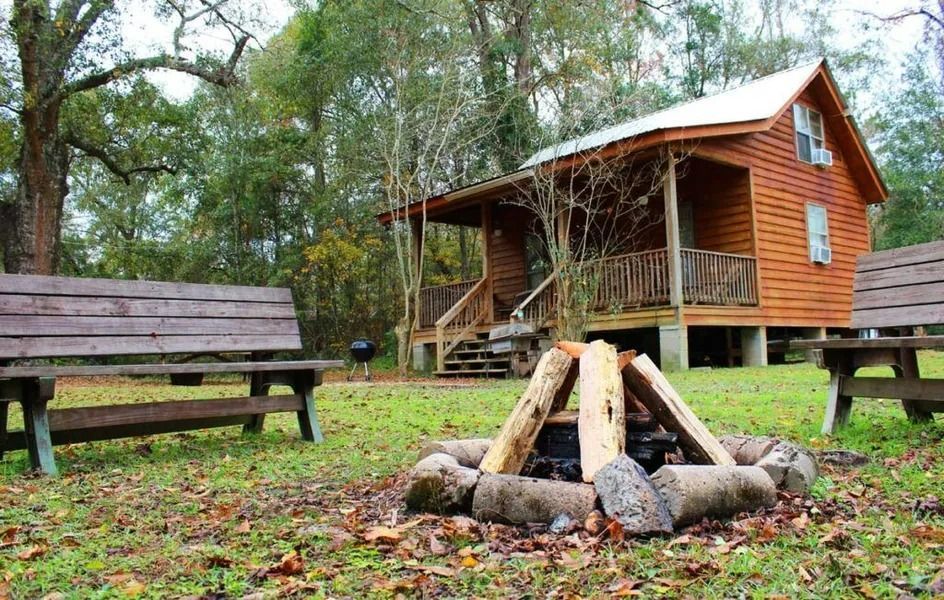 Wooden cabin in a forest clearing with benches and a campfire ring in the foreground