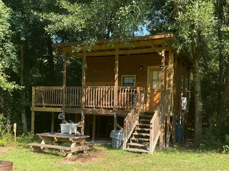 Small wooden cabin with porch and stairs in a wooded clearing, with a picnic table in front.