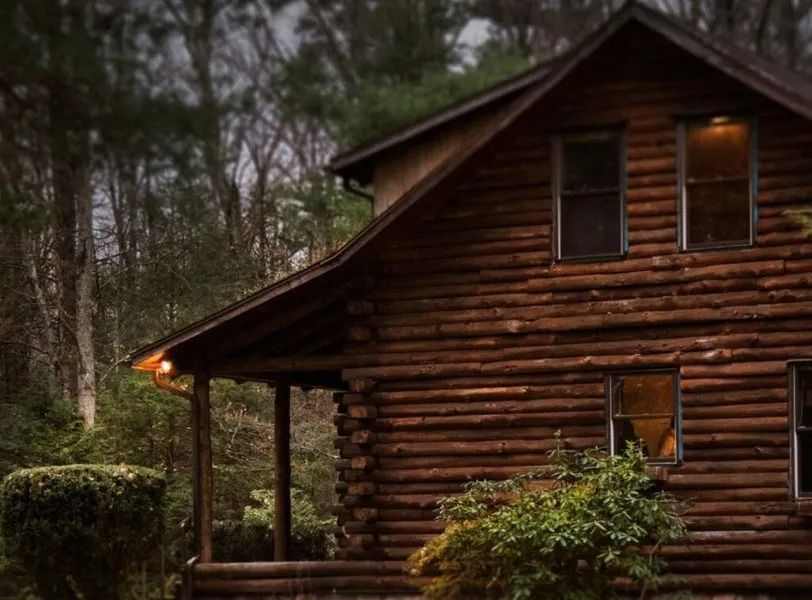 Rustic log cabin with a porch, nestled among trees in a wooded setting at dusk