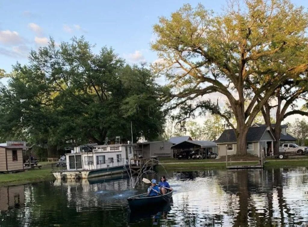 Rowboat on a calm river with a houseboat, trees, and houses along the shore at sunset