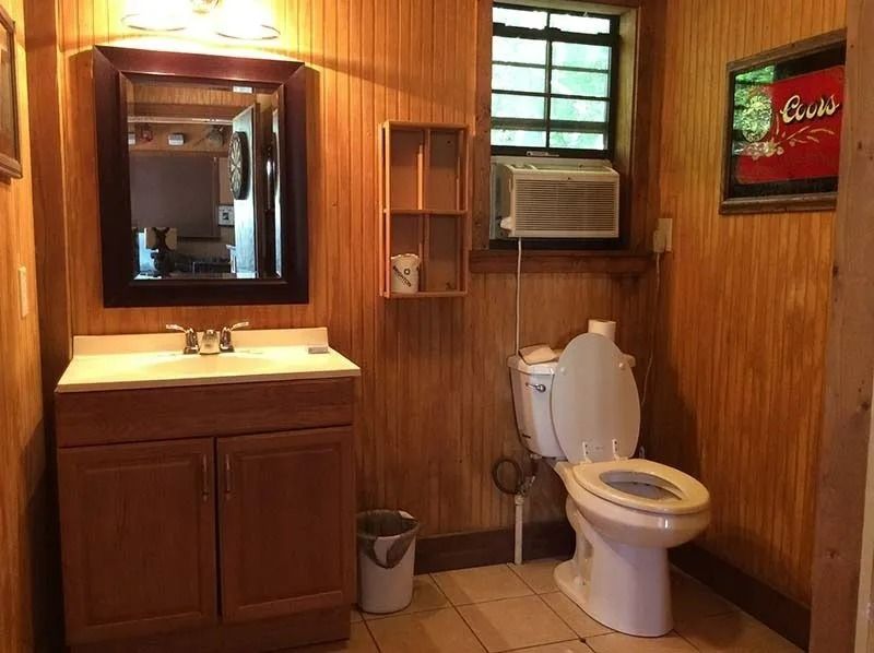 Small wood-paneled bathroom with sink, mirror, toilet, window AC unit, and trash can.